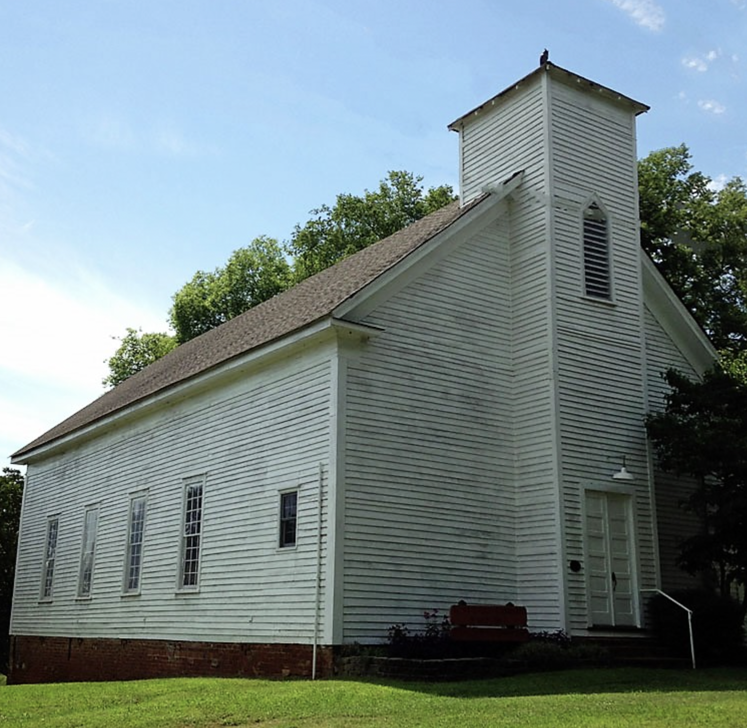 A white wooden church with a small steeple, surrounded by green grass and trees, under a blue sky.