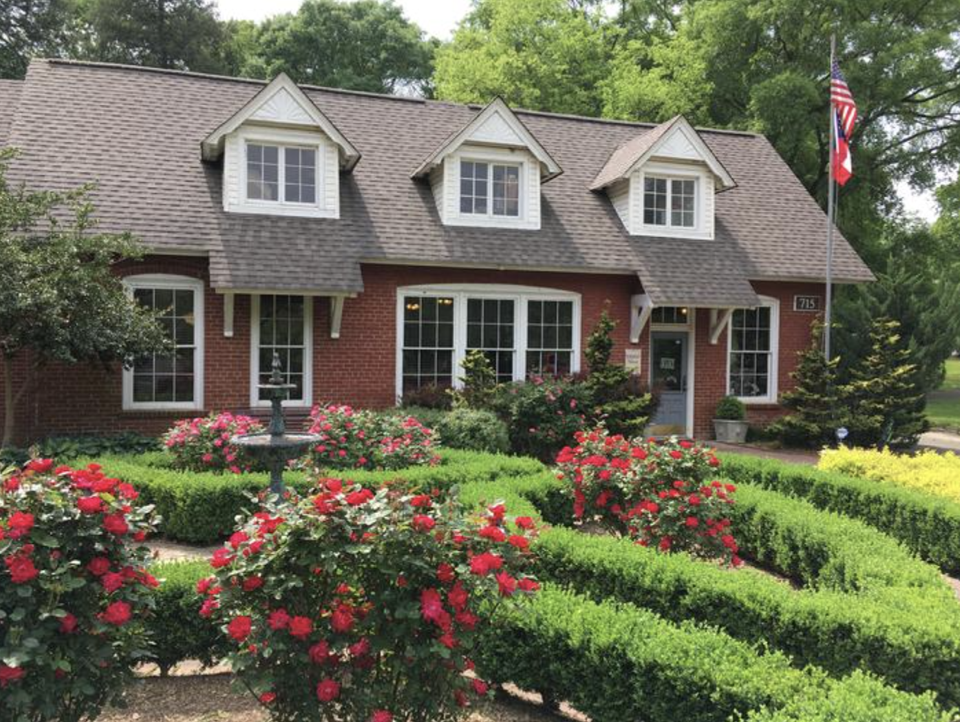 A brick house with a brown shingle roof and white window frames, surrounded by a well-maintained garden with blooming pink and red roses, green hedges, small trees, and an American flag flying on a pole.