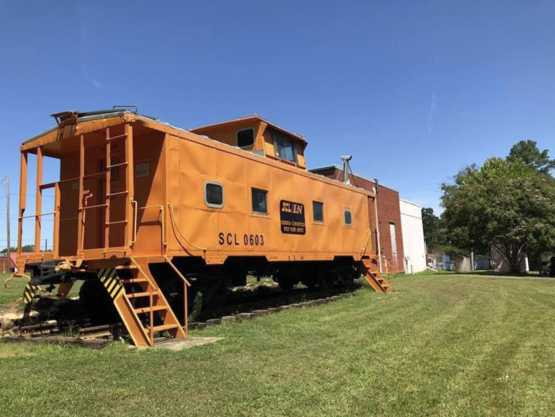 Orange caboose train car on grass with a building and trees in the background, under a clear blue sky.