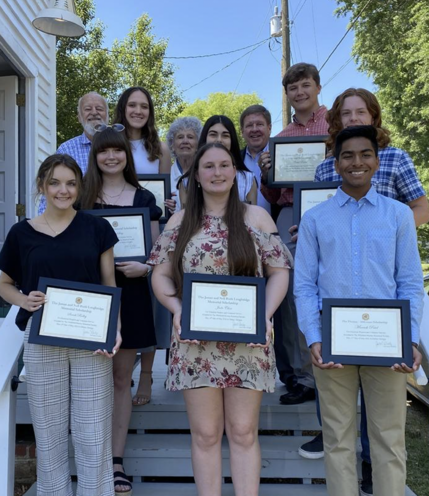 A group of young people and older adults standing on steps outside, holding framed certificates, smiling, during an award or graduation ceremony on a sunny day.