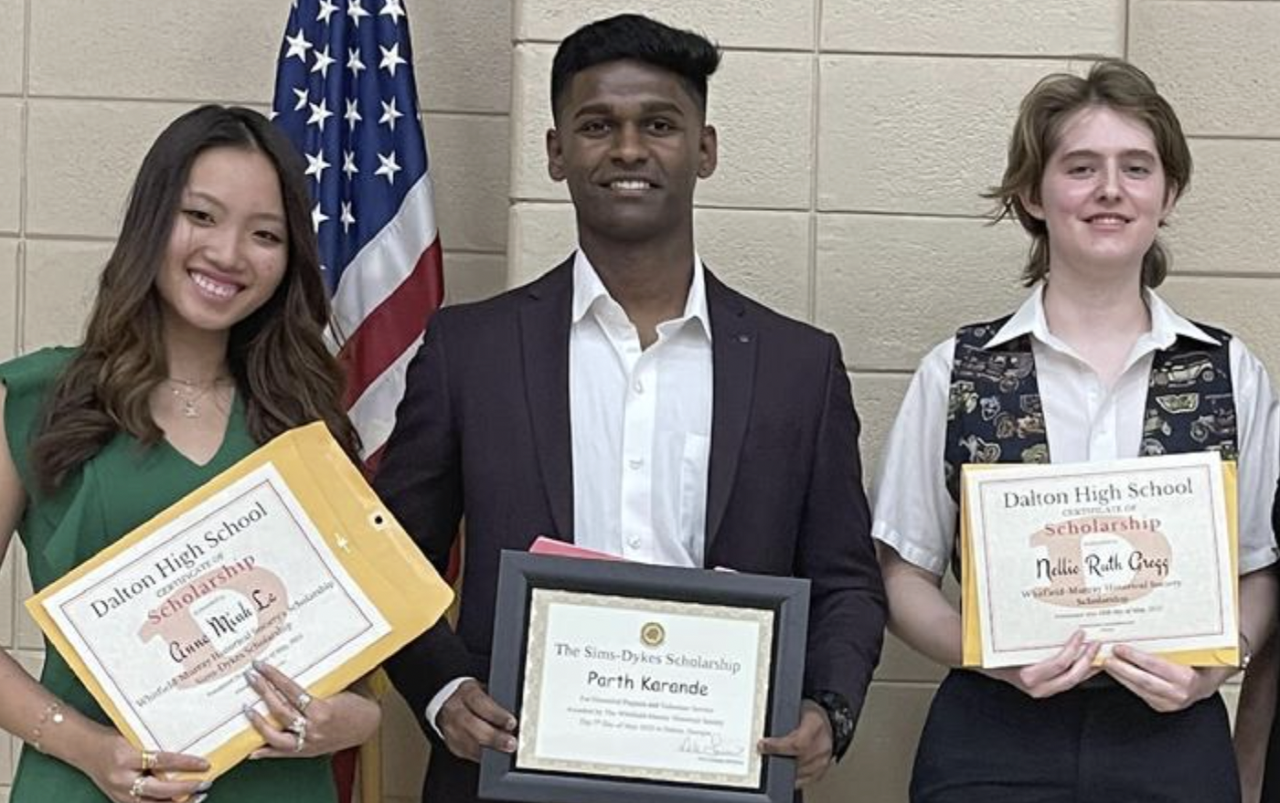 Three students receiving scholarships, two females holding certificates and one male holding a framed award, standing in front of an American flag and a beige wall.