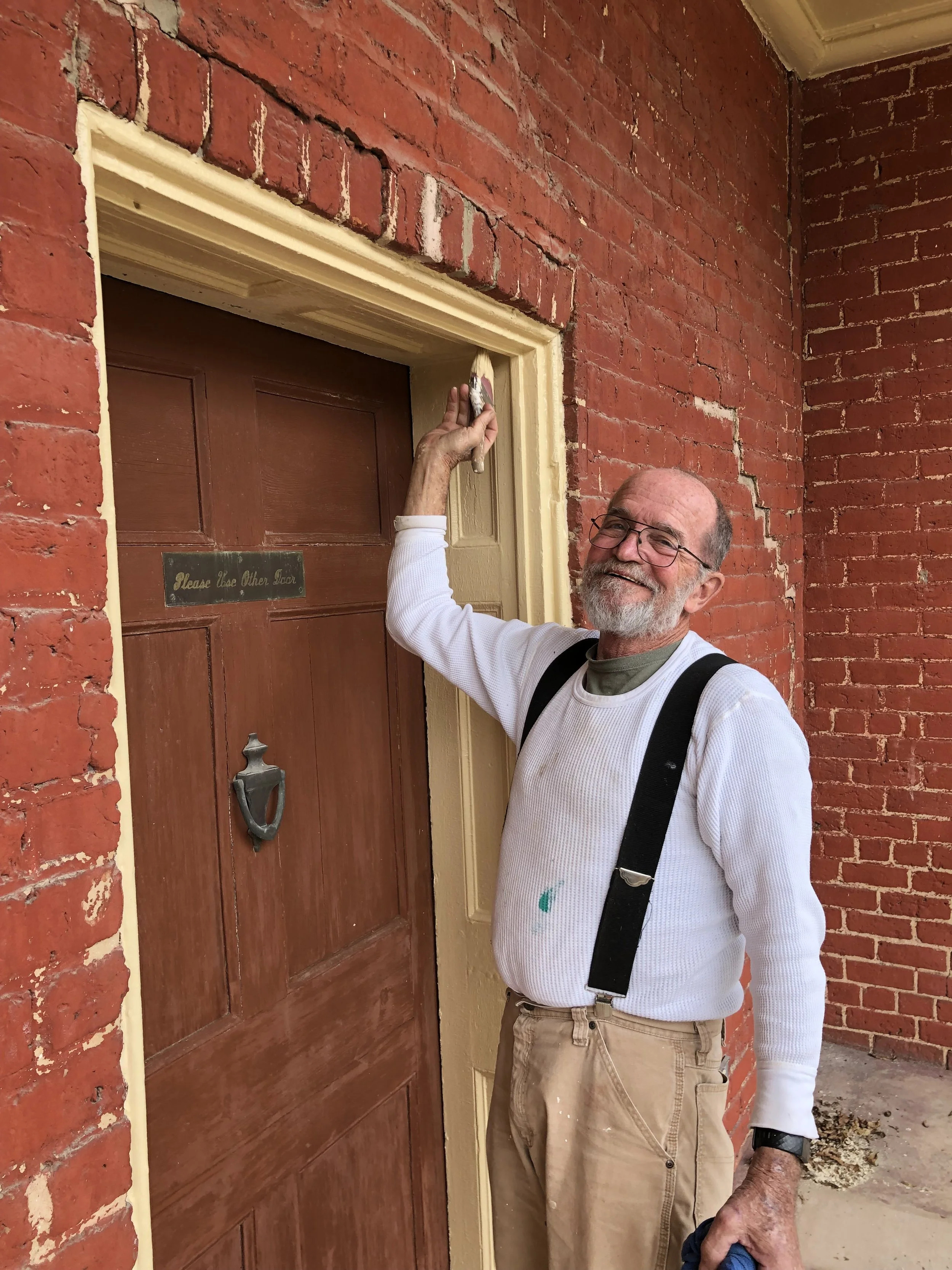 An older man with glasses, a beard, and a smile is painting the trim around a brown wooden door with beige paint. He is wearing a white long-sleeve shirt, suspenders, and beige pants. He is standing outside in front of a red brick wall and is holding a paintbrush in his right hand and cleaning a cloth in his left hand.