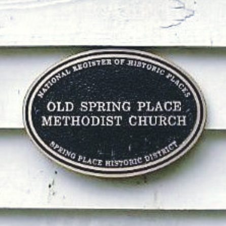 Close-up of a black oval sign with white text that reads 'Old Spring Place Methodist Church' and mentions it's part of the National Register of Historic Places and the Spring Place Historic District.