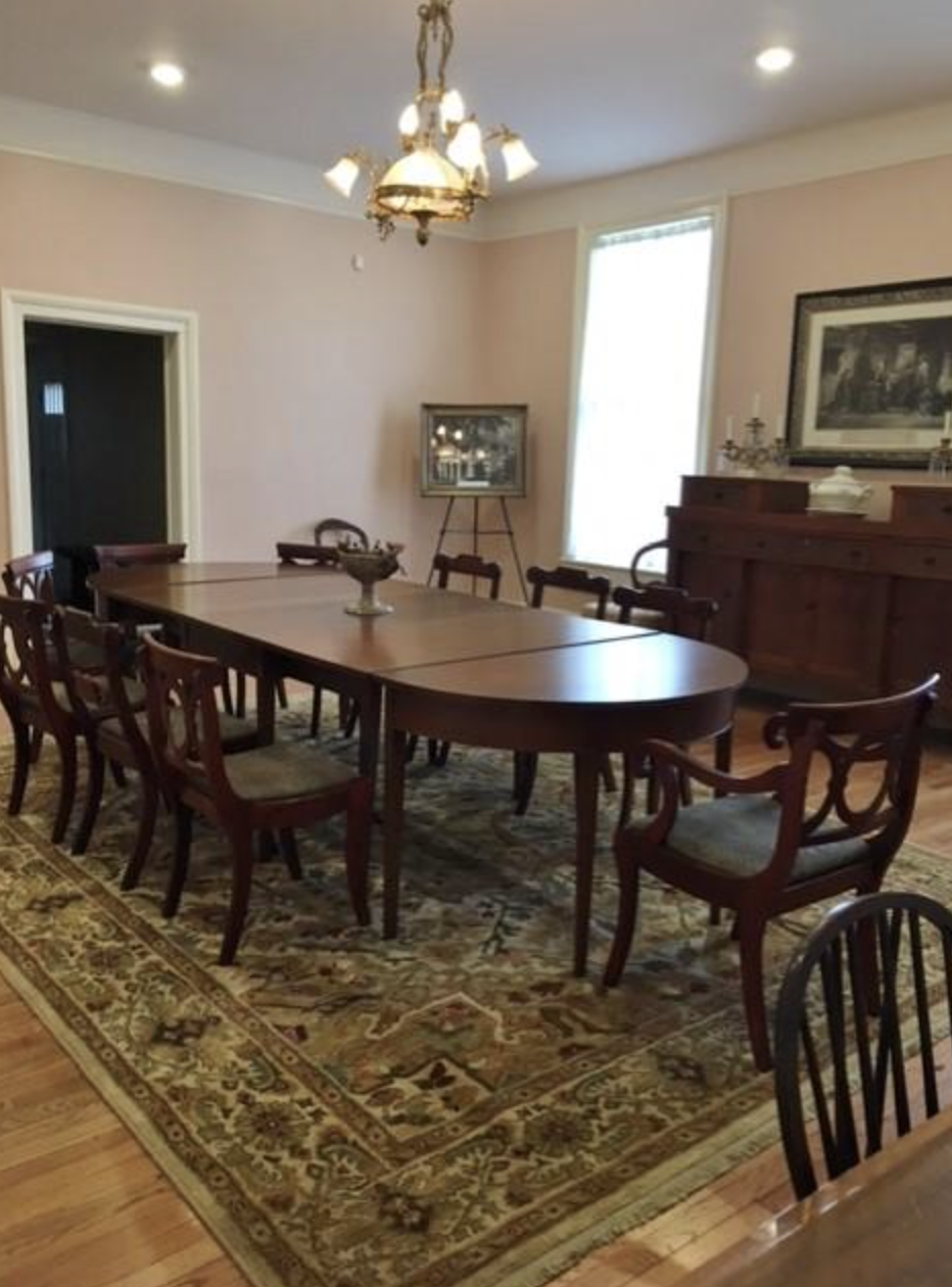 A formal dining room with a large wooden table, ten matching chairs, a patterned area rug, a chandelier, a sideboard with candles and artwork, a window with white curtains, and framed art on the walls.