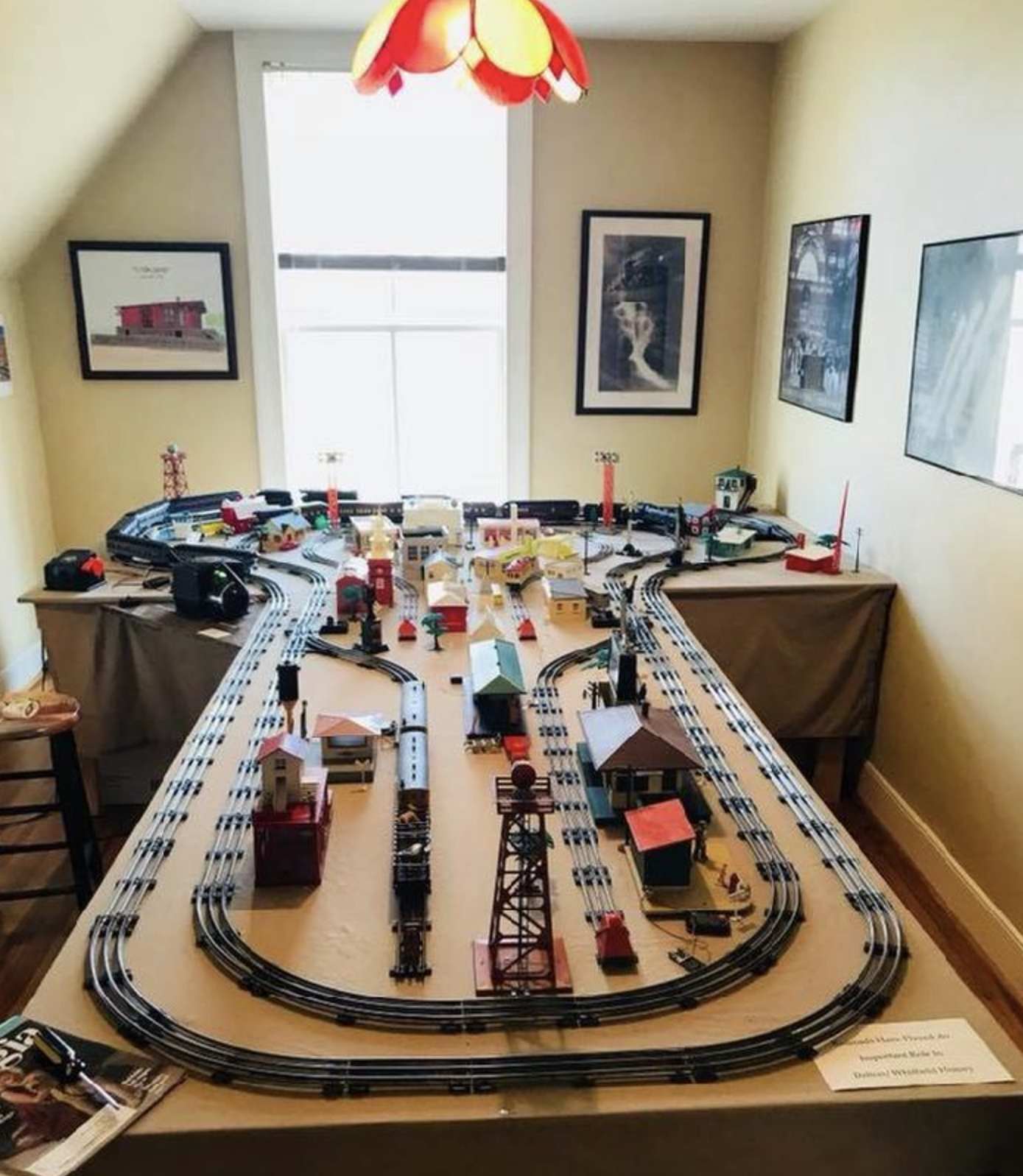 Model train set with tracks, buildings, and scenery on a long table in a room with yellow walls, framed pictures, and a red and yellow ceiling lamp.