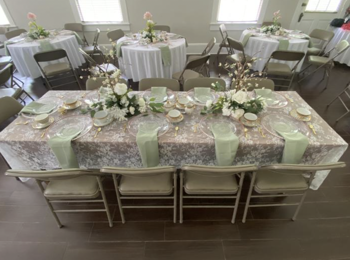 Elegant wedding reception table with a pink lace tablecloth, green napkins, delicate white and pink floral centerpieces, and gold-accented tableware, set in a bright room with multiple round tables and chairs.