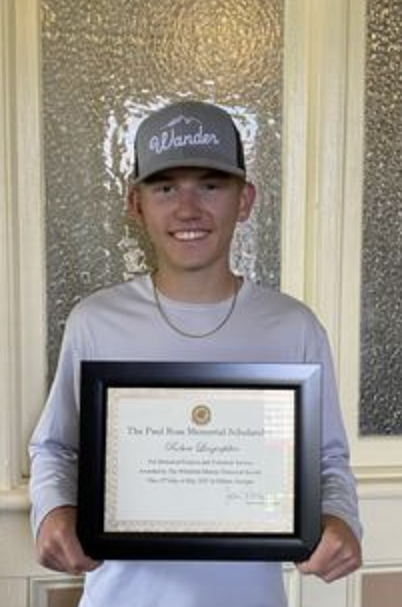 Young man wearing a gray and black baseball cap with 'Wander' written on it, holding a framed certificate, standing in front of textured glass doors.