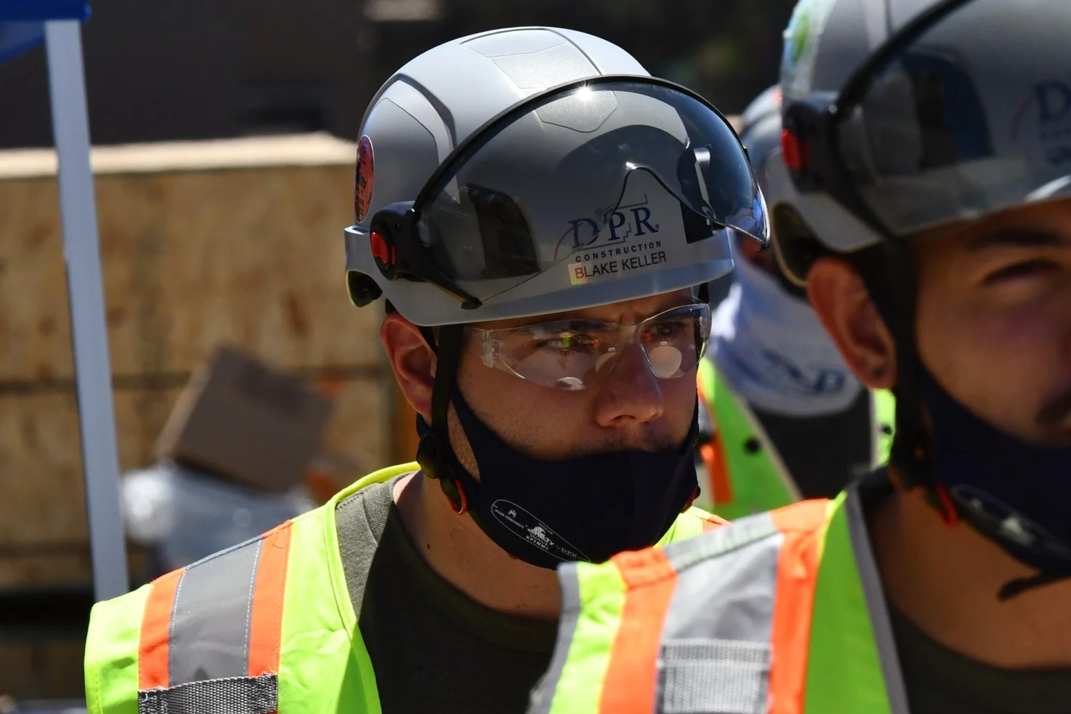 Construction workers wearing helmets, safety glasses, face masks, and high-visibility vests at a construction site.