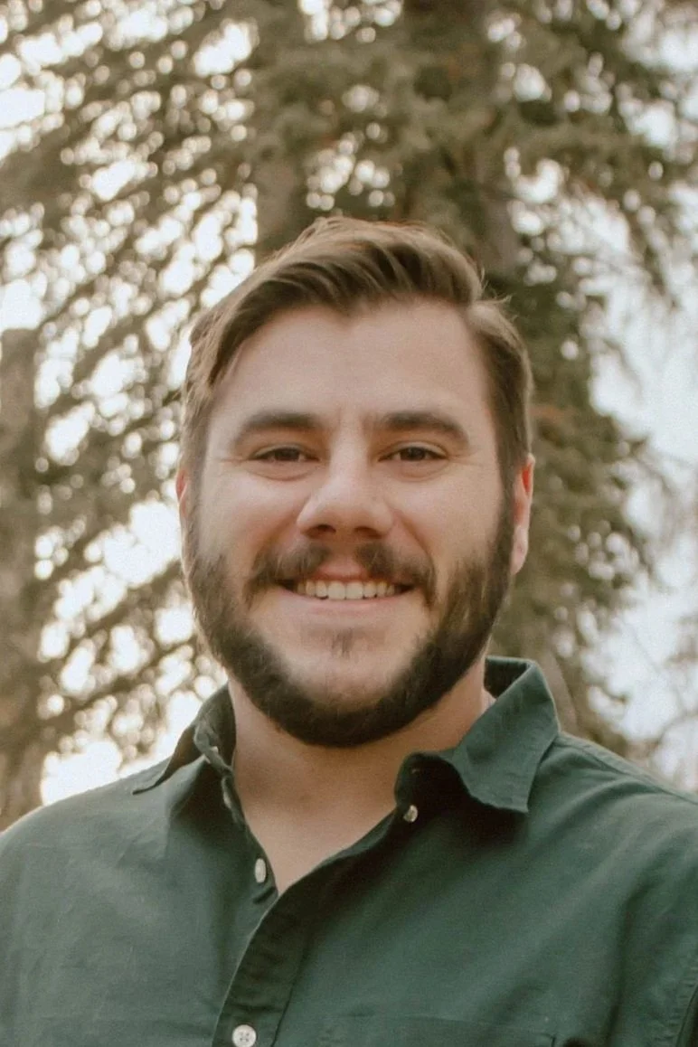 A man with a beard and short brown hair smiling outdoors, wearing a dark green button-up shirt, with trees in the background.