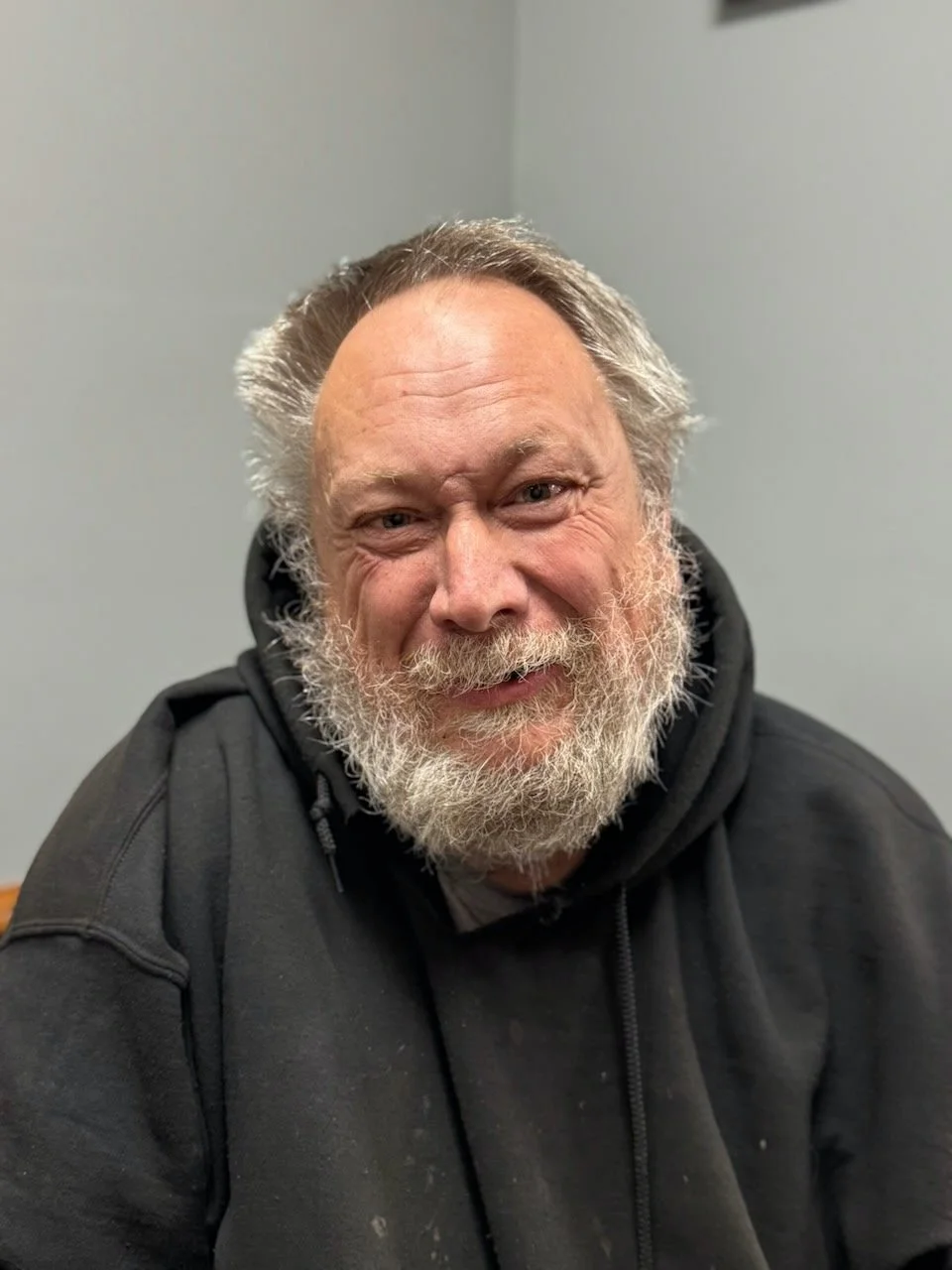A smiling middle-aged man with a full gray beard, gray hair, and wearing a black hoodie, sitting in a room with a plain, light-colored wall.