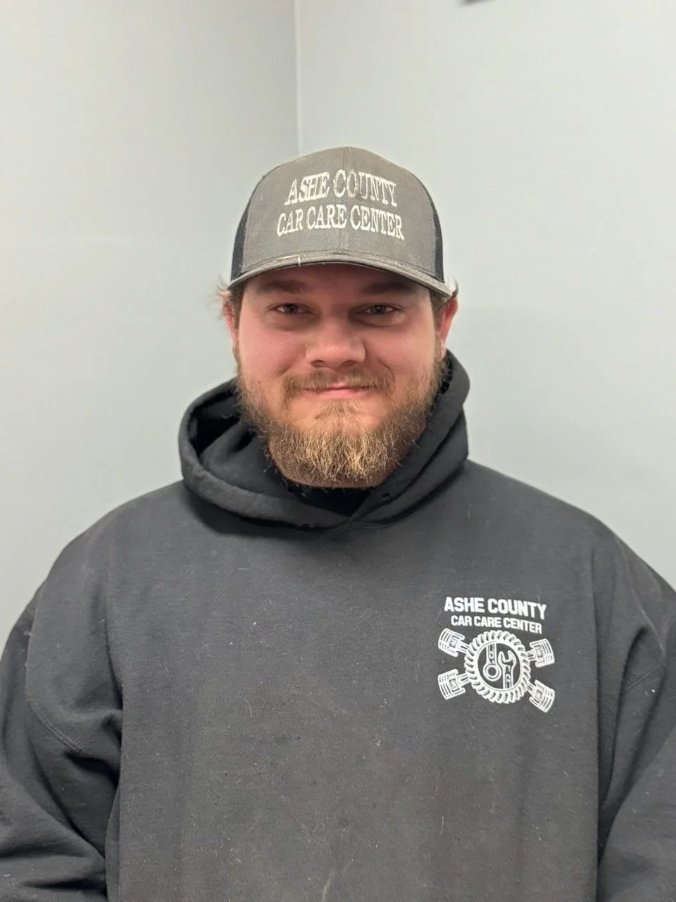A man with a beard wearing a charcoal gray hat and hoodie from Ashe County Car Care Center, standing in front of a plain light-colored wall.