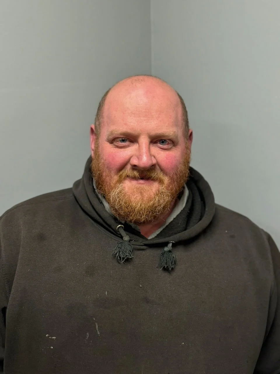 A smiling man with a red beard and short hair, wearing a dark hoodie, against a plain light gray wall.