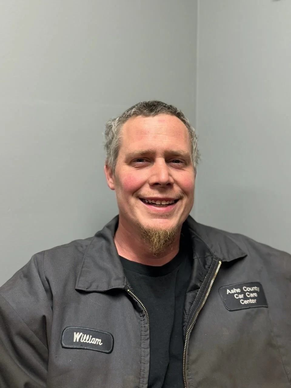 Smiling man in black uniform with name tag 'William' and 'Ashe County Car Care Center' patch, inside a plain gray room.