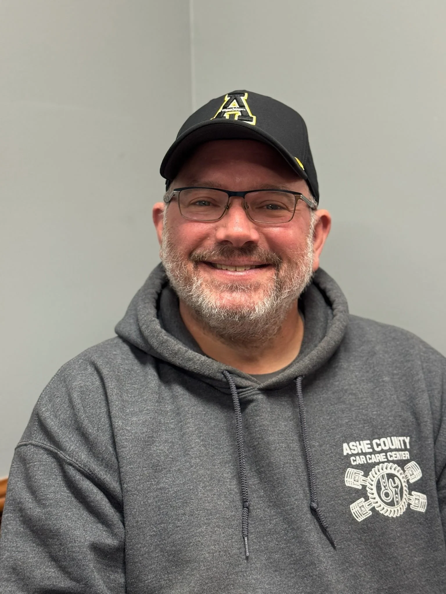 A smiling man with glasses, a beard, and mustache, wearing a black baseball cap and a gray hoodie with 'Ashe County Car Care Center' logo, sitting indoors against a plain gray wall.
