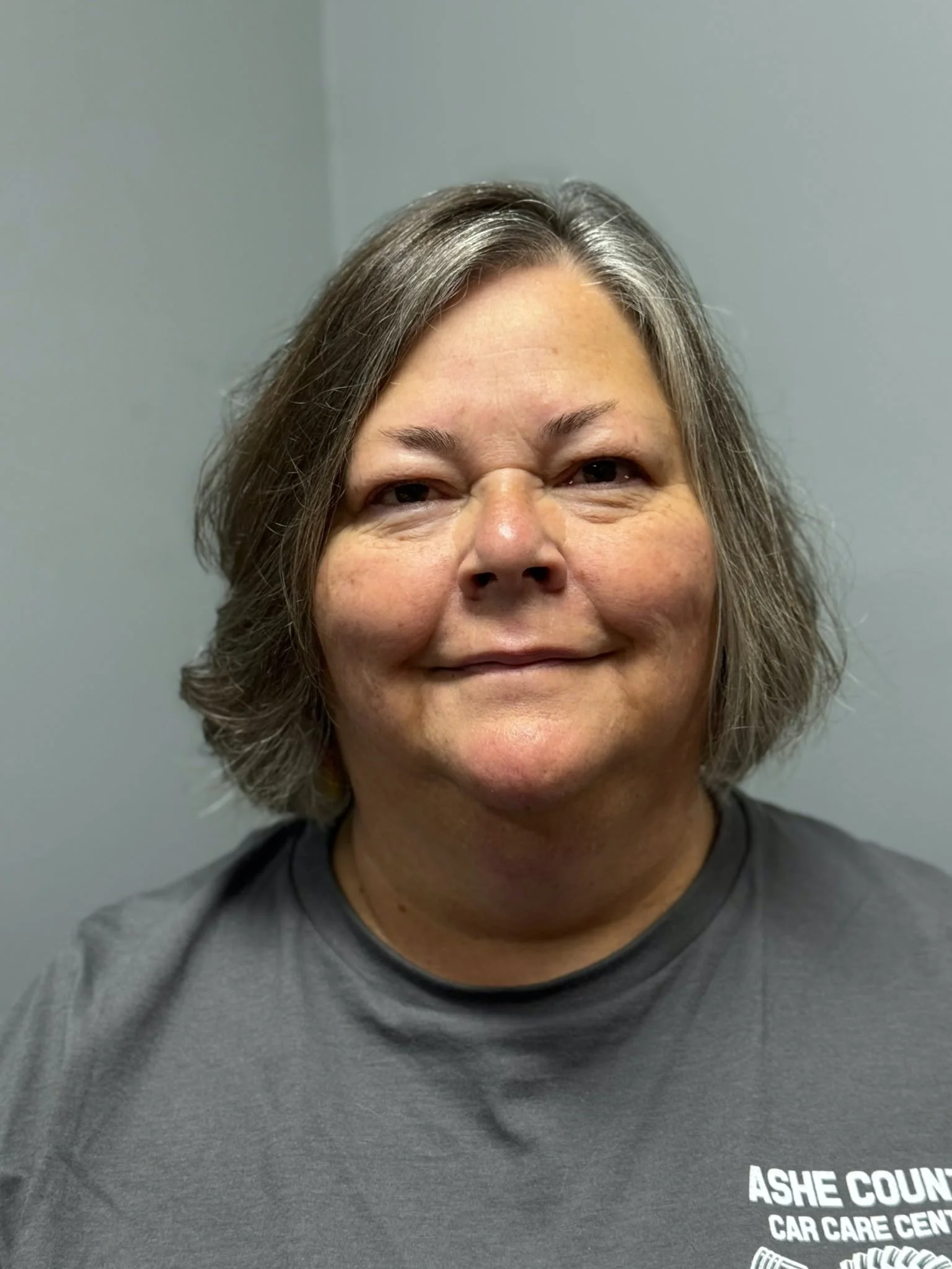 A middle-aged woman with shoulder-length gray hair, smiling, wearing a dark gray T-shirt with white text that reads "ASHE COUNT CAR CARE CENT" against a plain gray background.