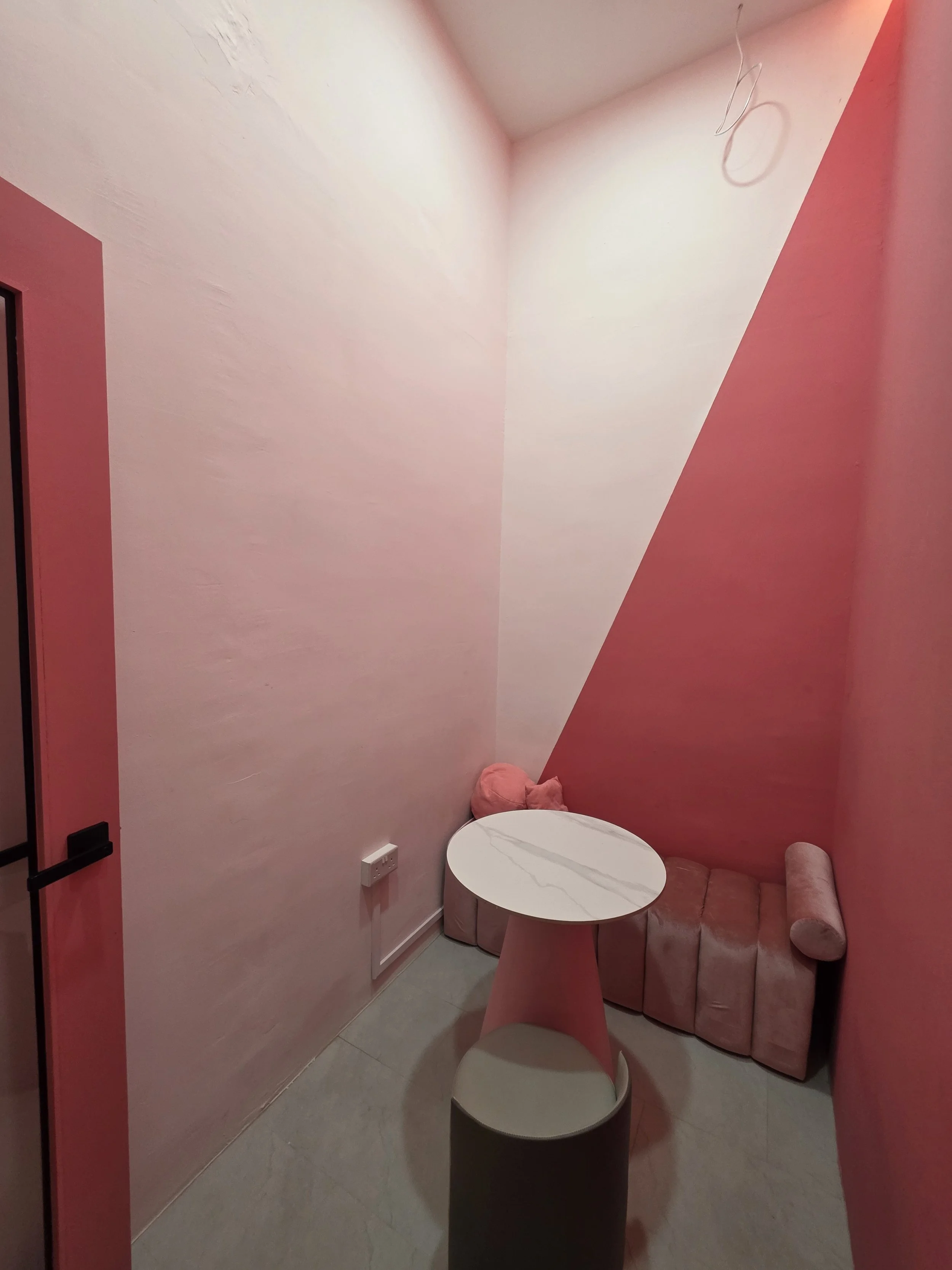 The Space Grid - Pink-colored corner with angled walls, a small round white marble table, pink plush cushions, a pink upholstered bench, and an electrical outlet on the wall.