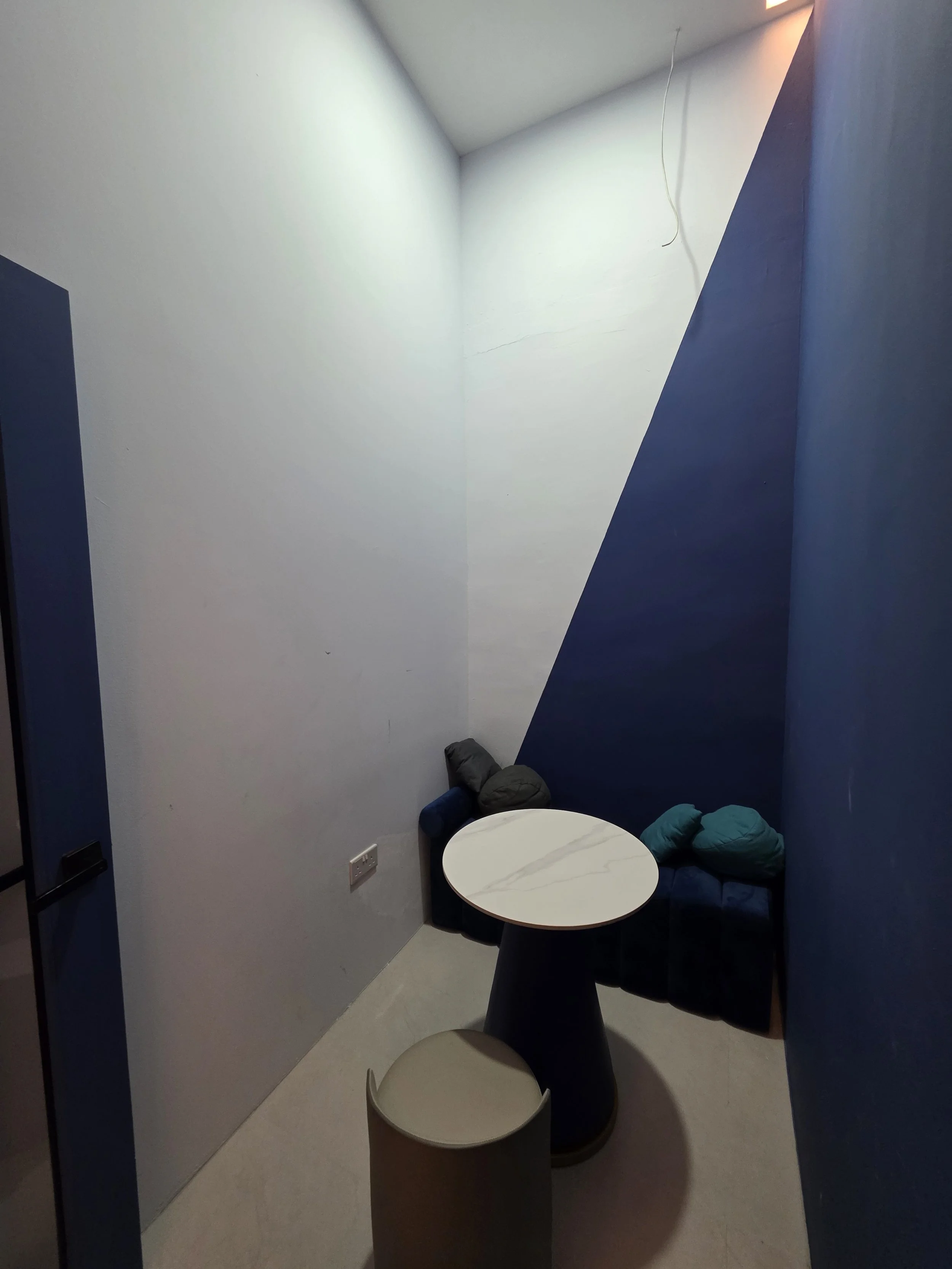 The Space Grid - Small corner seating area with a round table, two blue cushions, and a blue padded bench against the wall. The walls are painted in white and dark blue geometric patterns. There is a black cylindrical stool in front of the table.