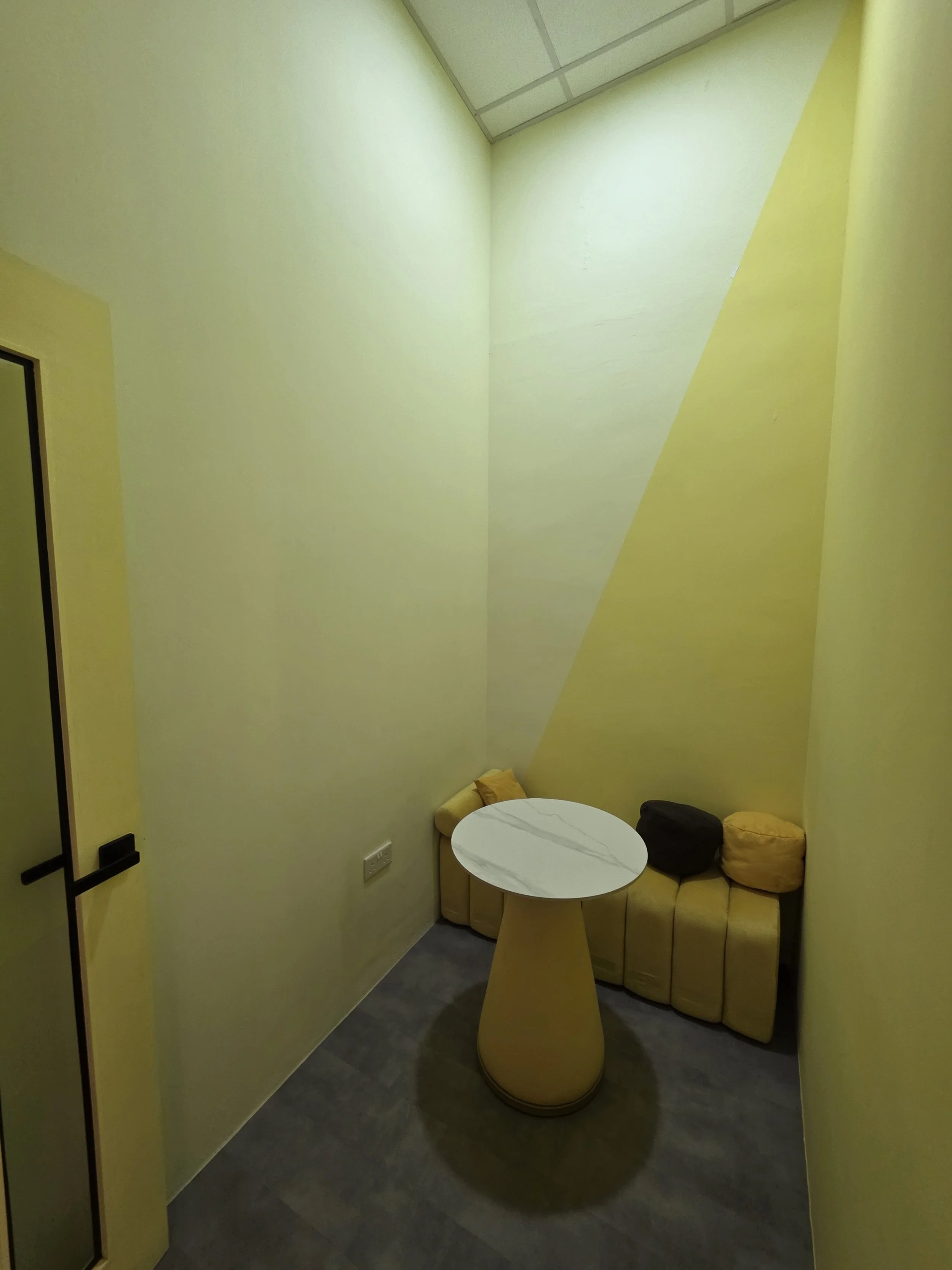 The Space Grid - Small yellow corner seating area with a yellow upholstered bench, two yellow cushions, a black cushion, a small yellow side table, and yellow walls with a geometric design.