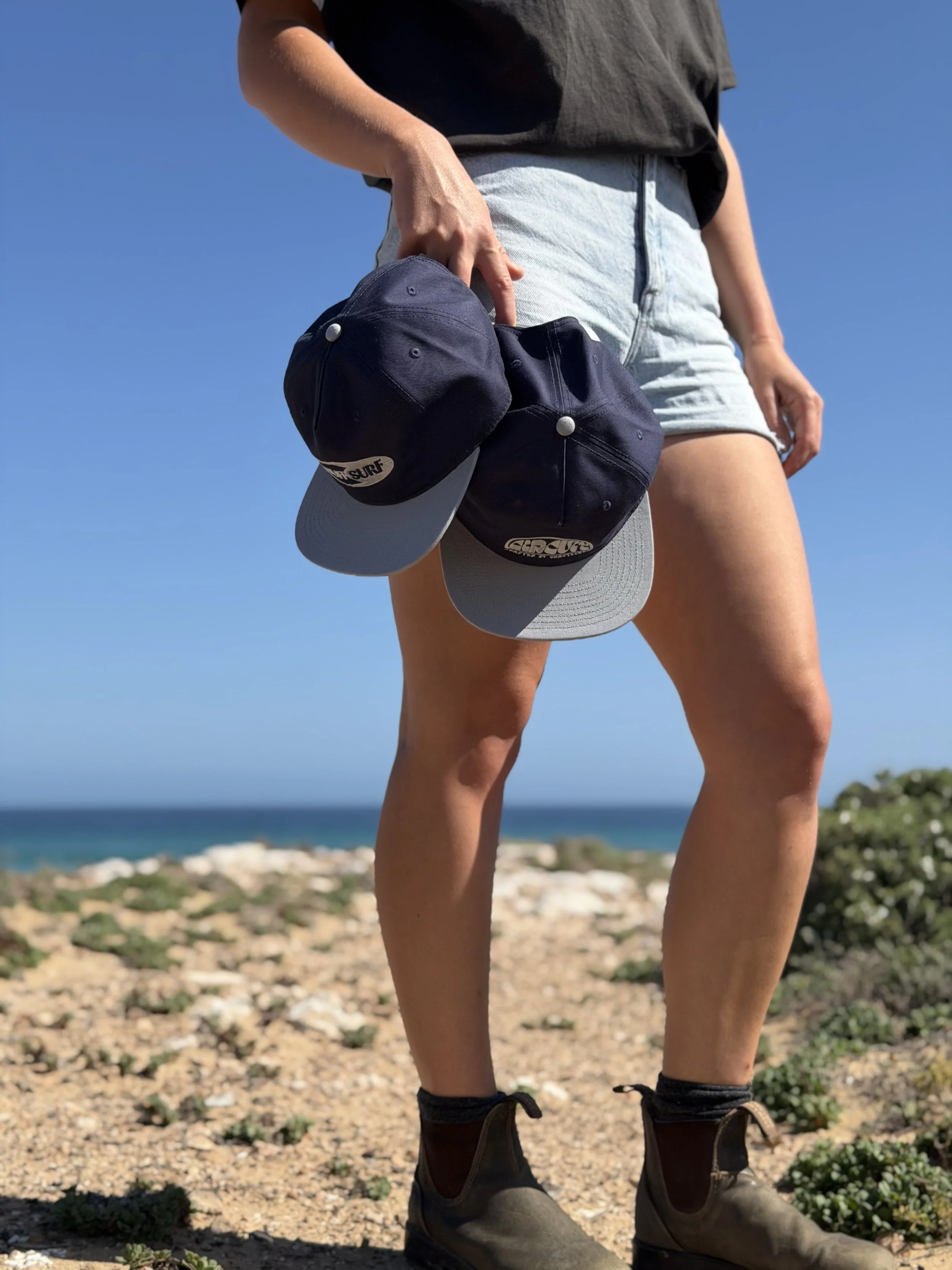 A person standing outdoors on rocky terrain near the coast, holding a dark baseball cap with a white brim in their hand, with the ocean and a clear blue sky in the background.