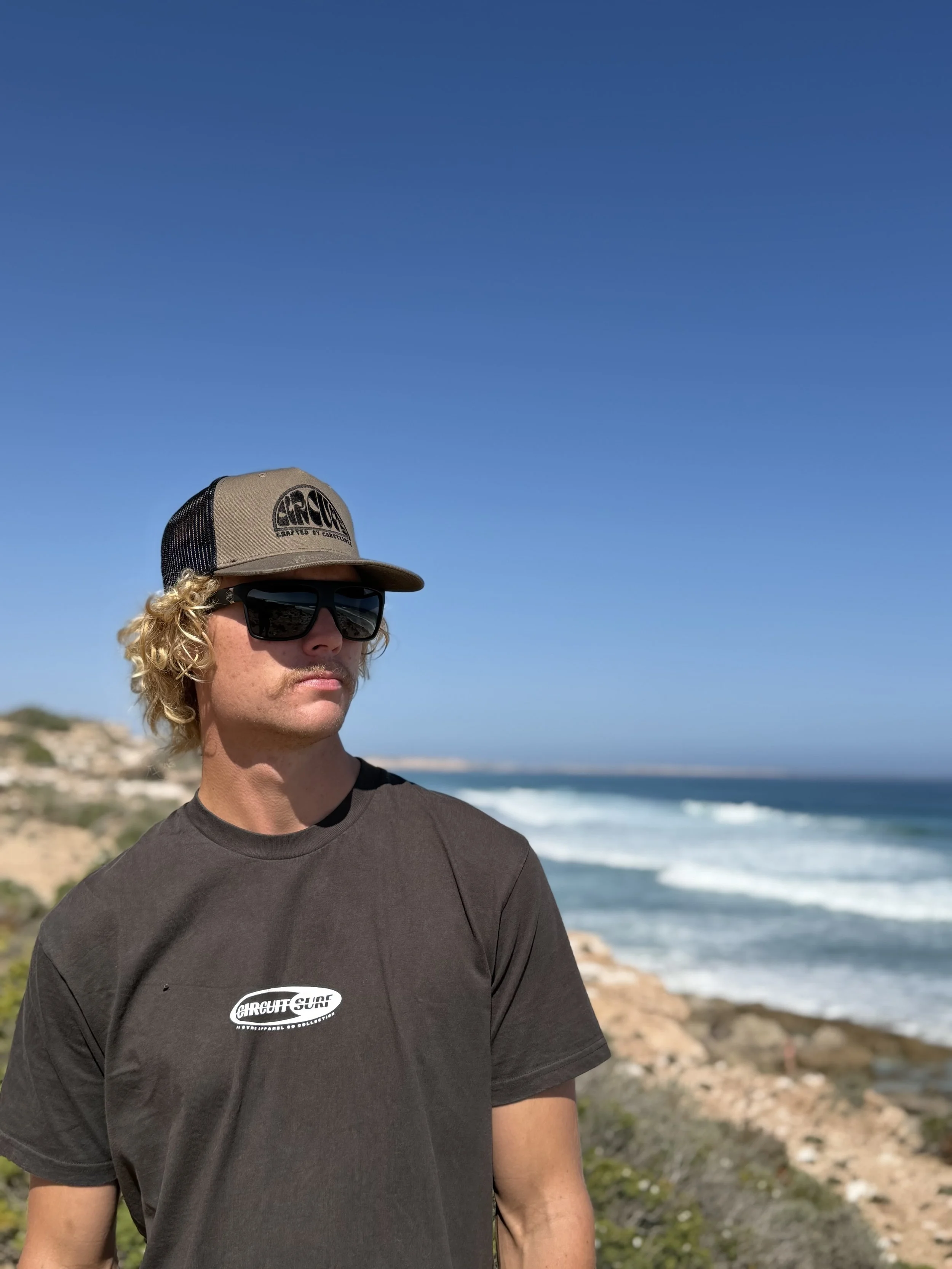 A person with curly blonde hair, wearing a gray cap and sunglasses, standing on a rocky beach with the ocean and clear blue sky in the background.