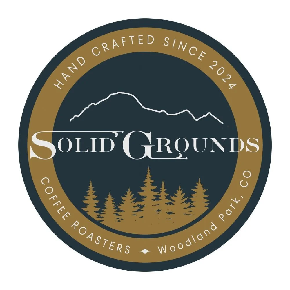 Circular logo for Solid Grounds Coffee Roasters, featuring a mountain outline at the top, a forest of pine trees at the bottom, and text indicating it has been handcrafted since 2024, located in Woodland Park, CO.