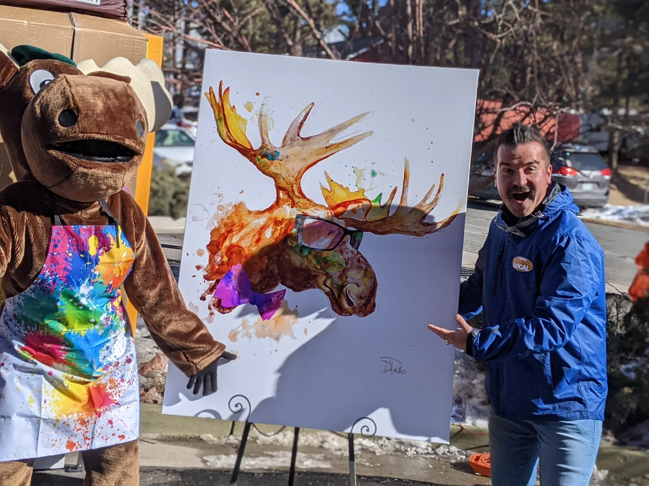 A person in a moose costume and a man are outdoors, displaying a colorful abstract painting of a moose on an easel. The man appears excited, and the background includes parked cars and trees.