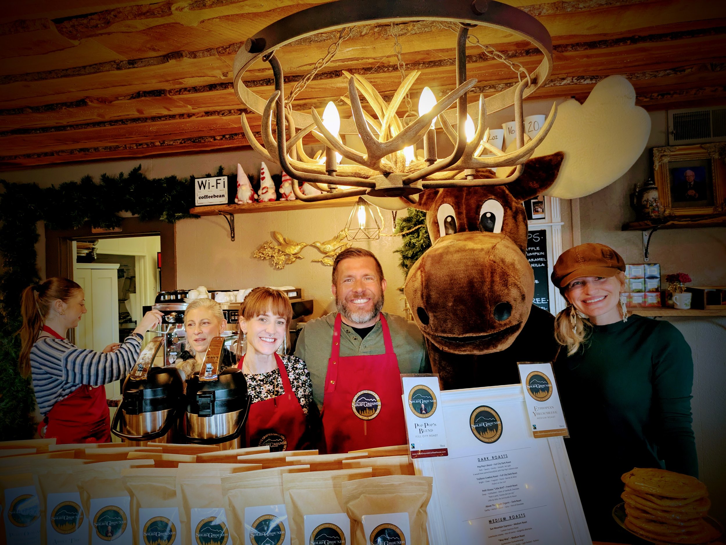 Group of five people, including two women, a man, and two individuals in a moose costume, standing behind a counter at a coffee shop with baked goods and coffee brewing equipment, festive decorations, and a moose head decoration in the background.