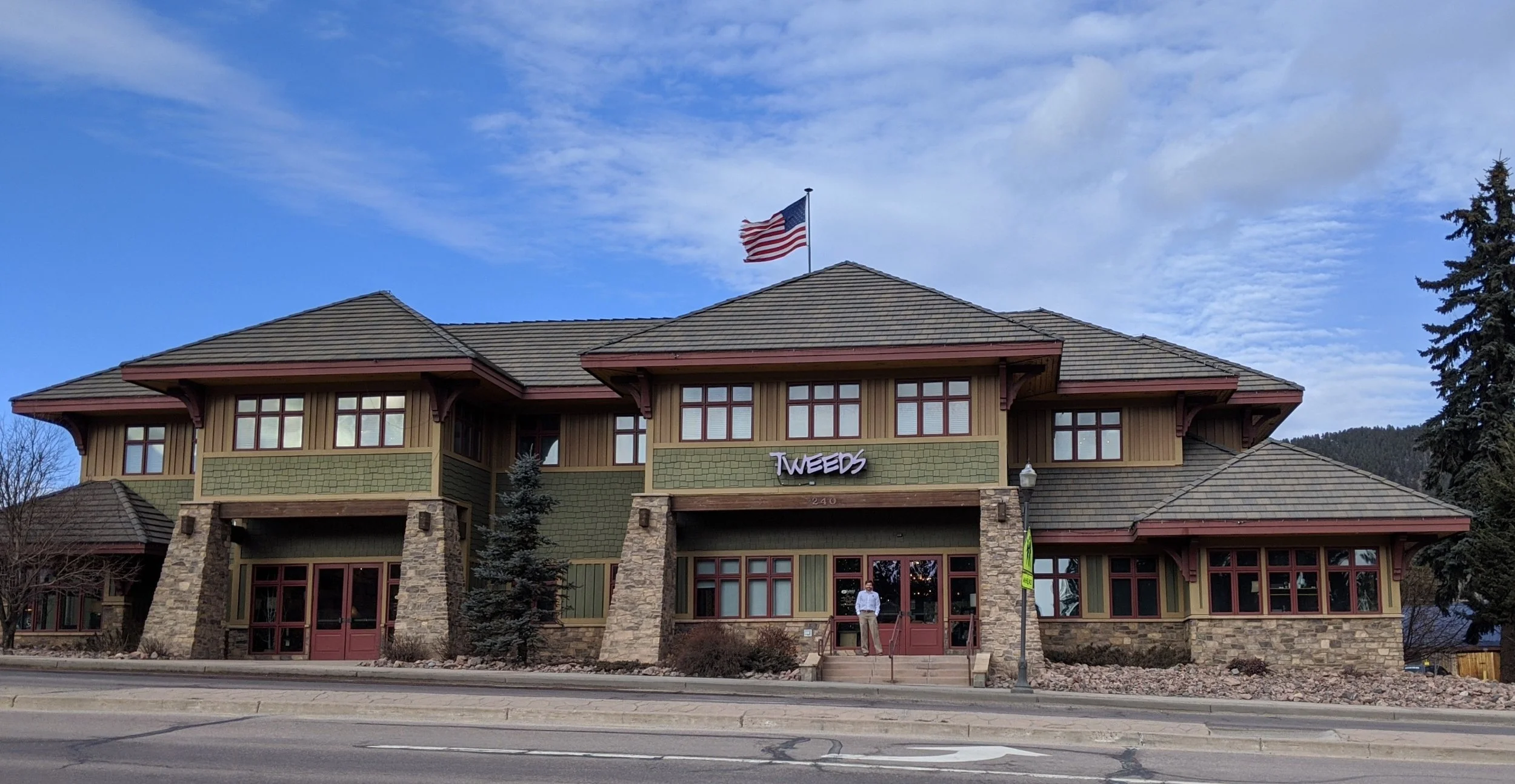 A two-story building with green and brown exterior, stone accents, and a sign that reads "TWEEDS" above the entrance. An American flag is flying on top of the building.