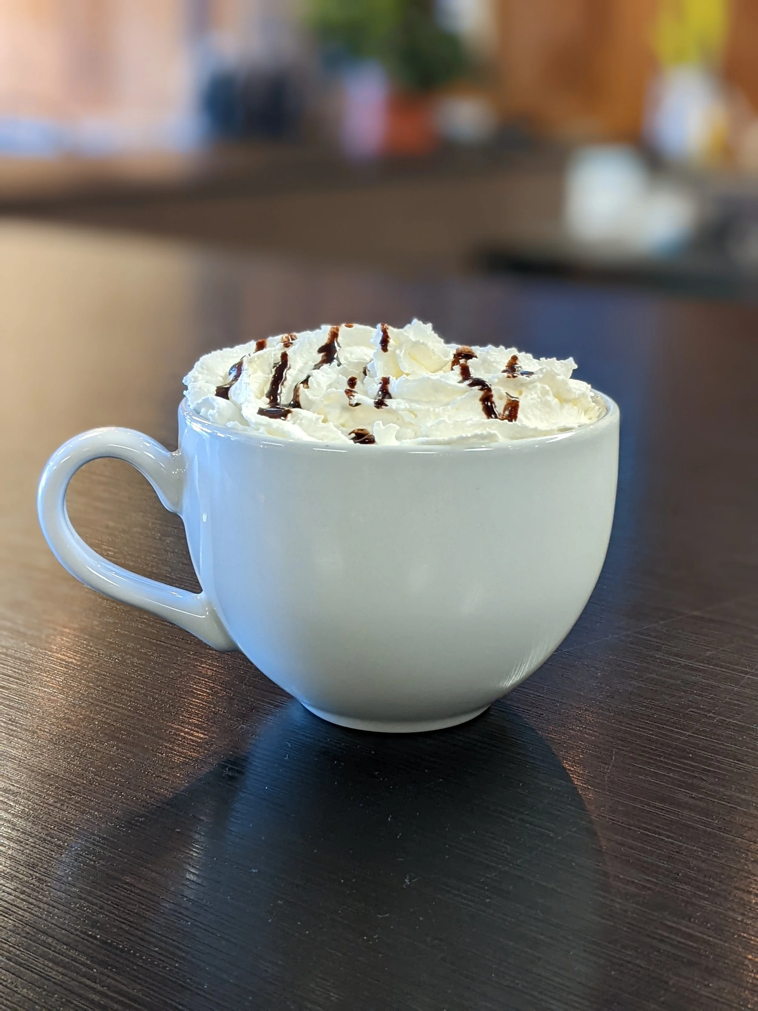 A white ceramic mug filled with a hot beverage topped with whipped cream and drizzled with chocolate syrup, placed on a dark wooden table.