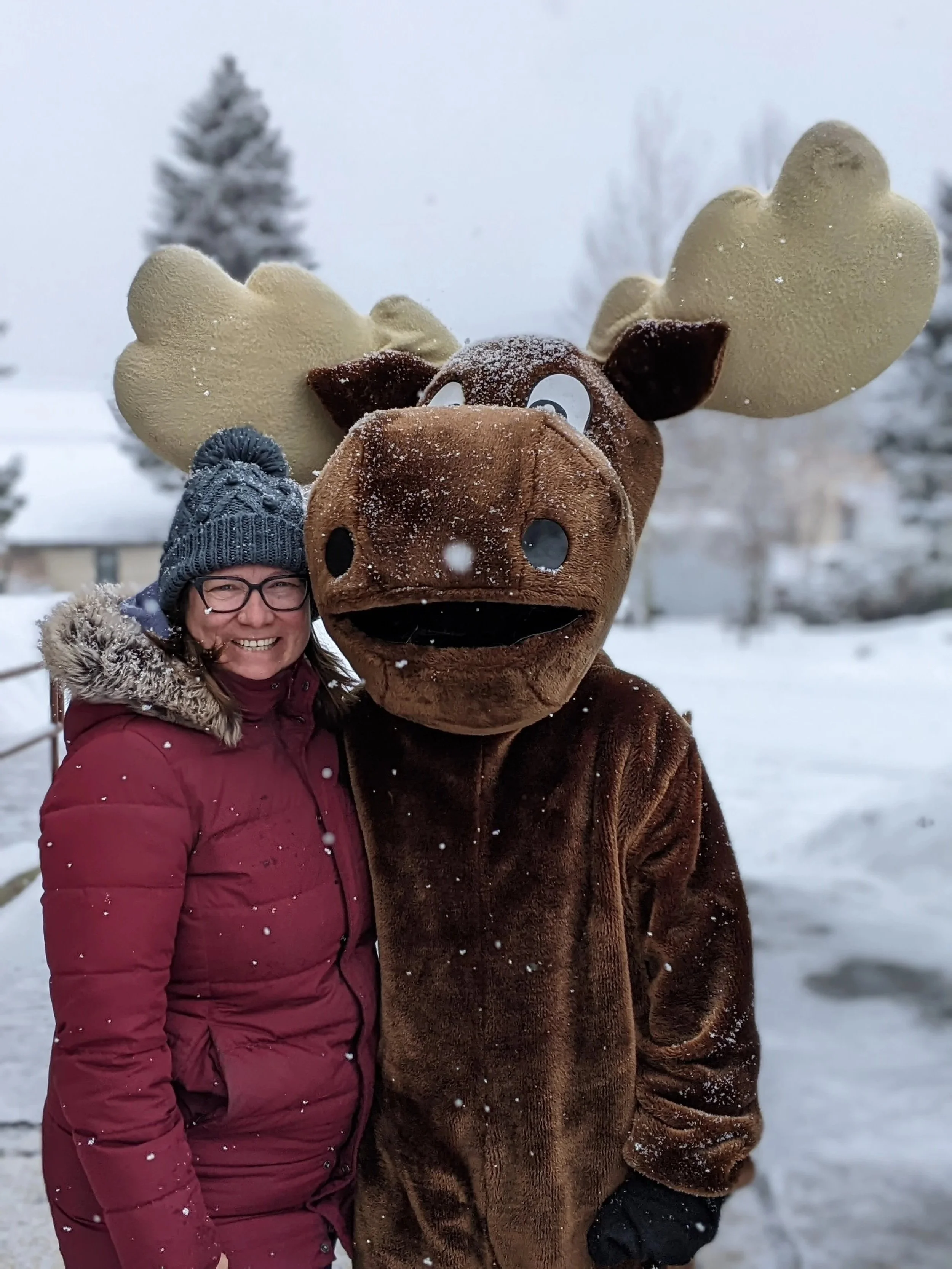 A woman in winter clothing and glasses standing next to a person in a moose costume outdoors in a snowy landscape.