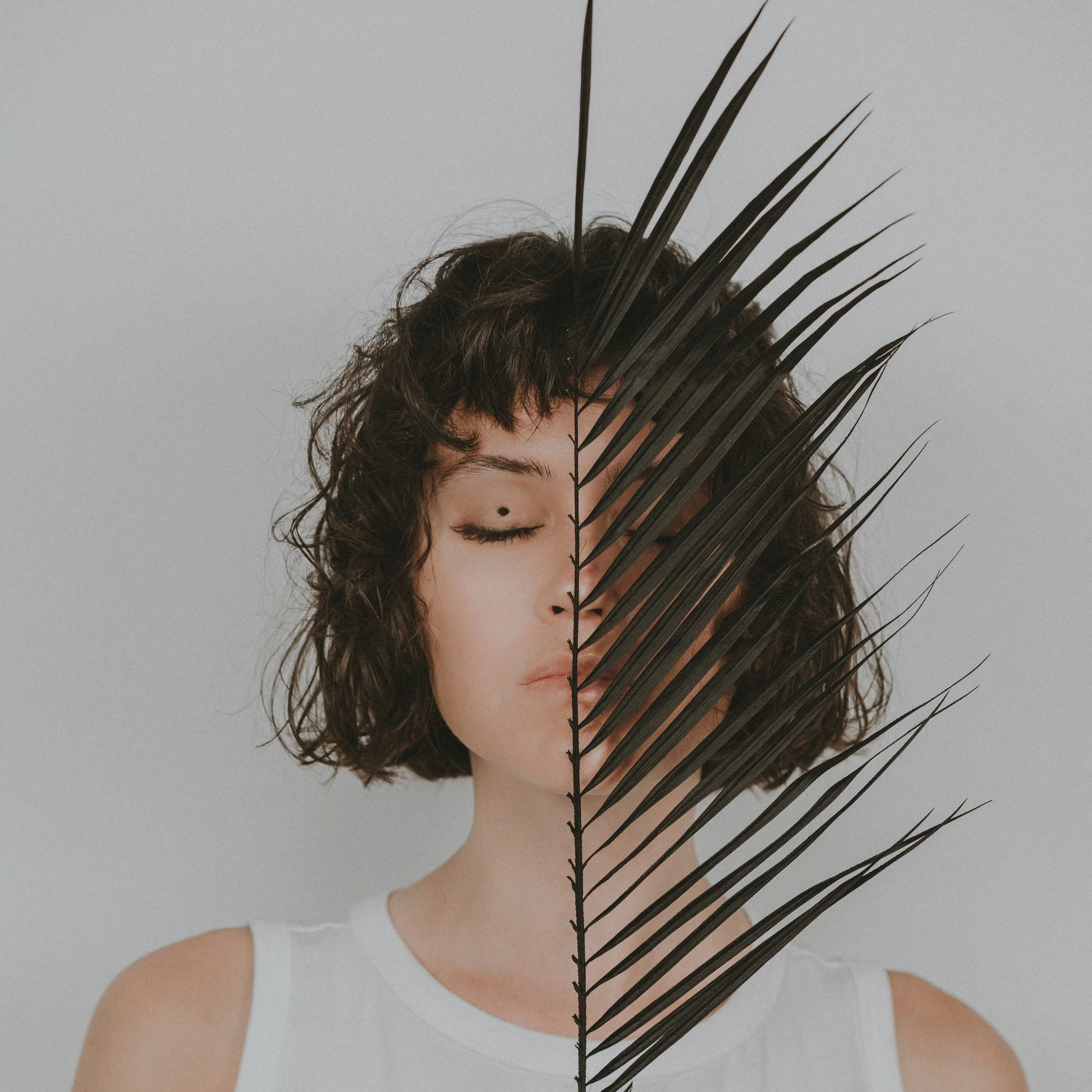 A woman with short, curly dark hair and closed eyes, wearing a white sleeveless top, is partially obscured by a large, dark palm leaf.