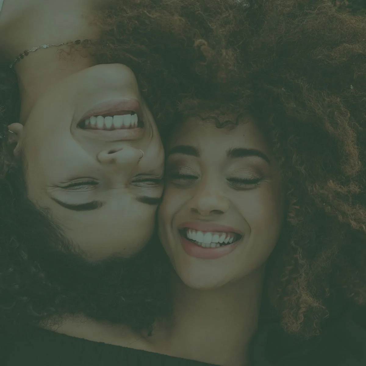Two women with curly hair lying on the ground close together, both smiling with their eyes closed.