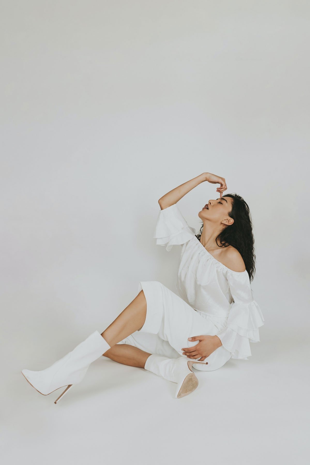 A woman in a white off-shoulder dress and white high-heeled boots sitting on the floor against a plain white background, posing with one hand on her head and the other on her stomach.