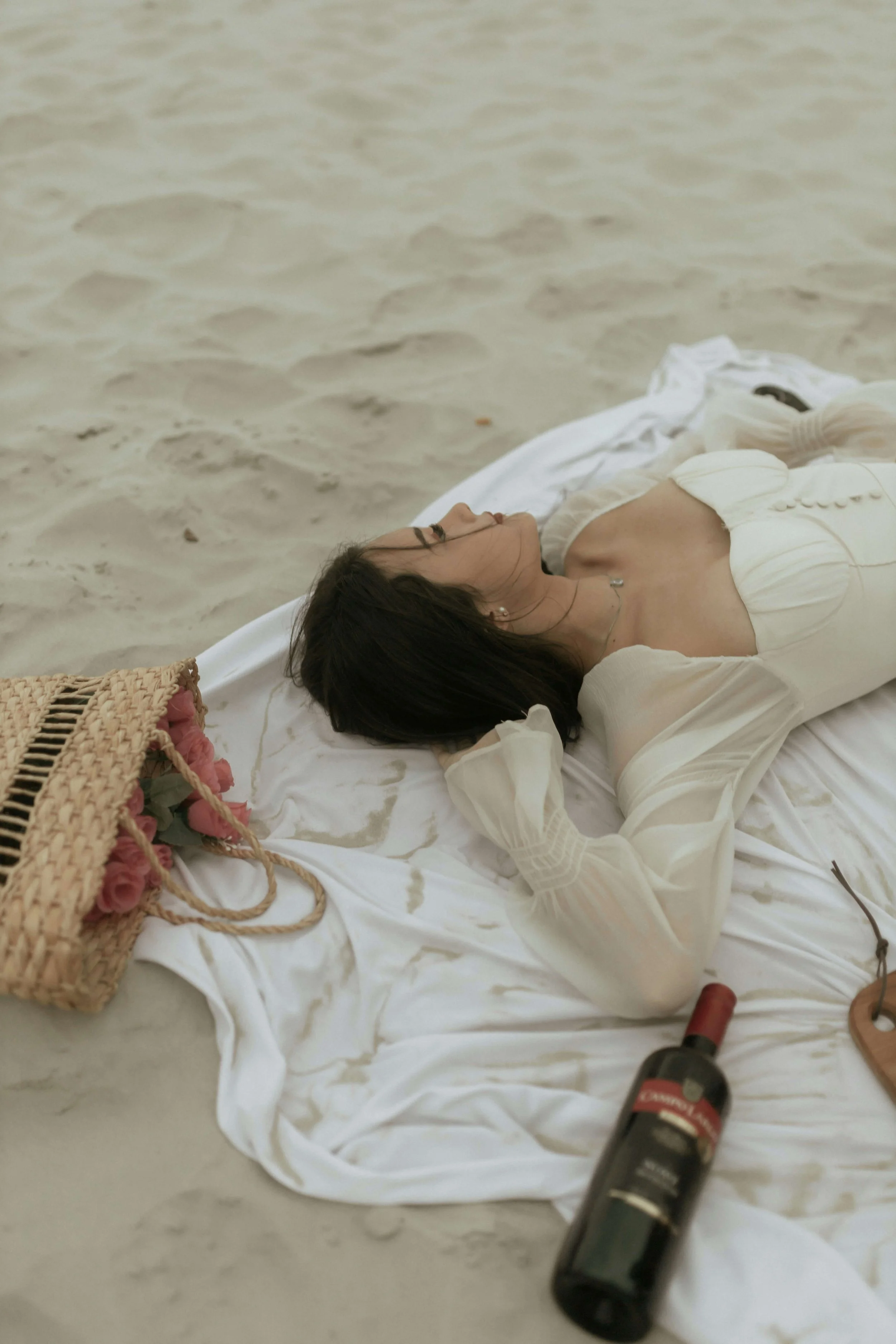 A woman lying on her back on a white blanket on the sand at the beach, relaxed and slowing down