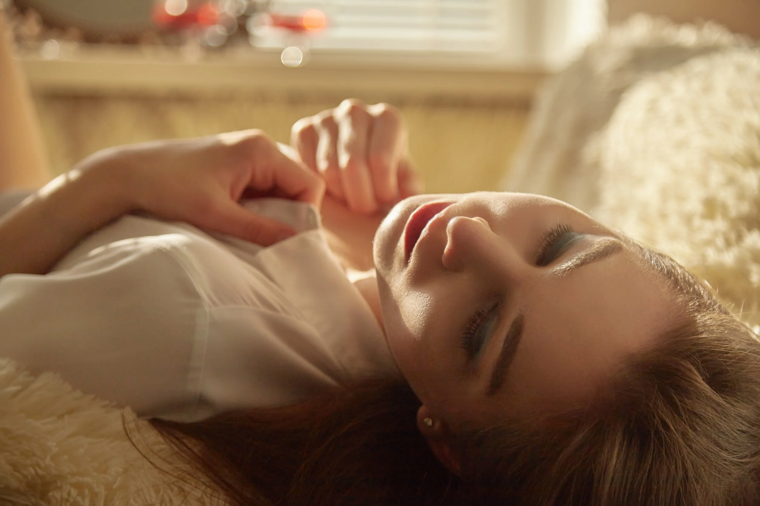 A woman with light skin and makeup lying on her back on a soft surface, with her eyes closed and hand resting on her chest, in a warmly lit room, feeling all the sensations
