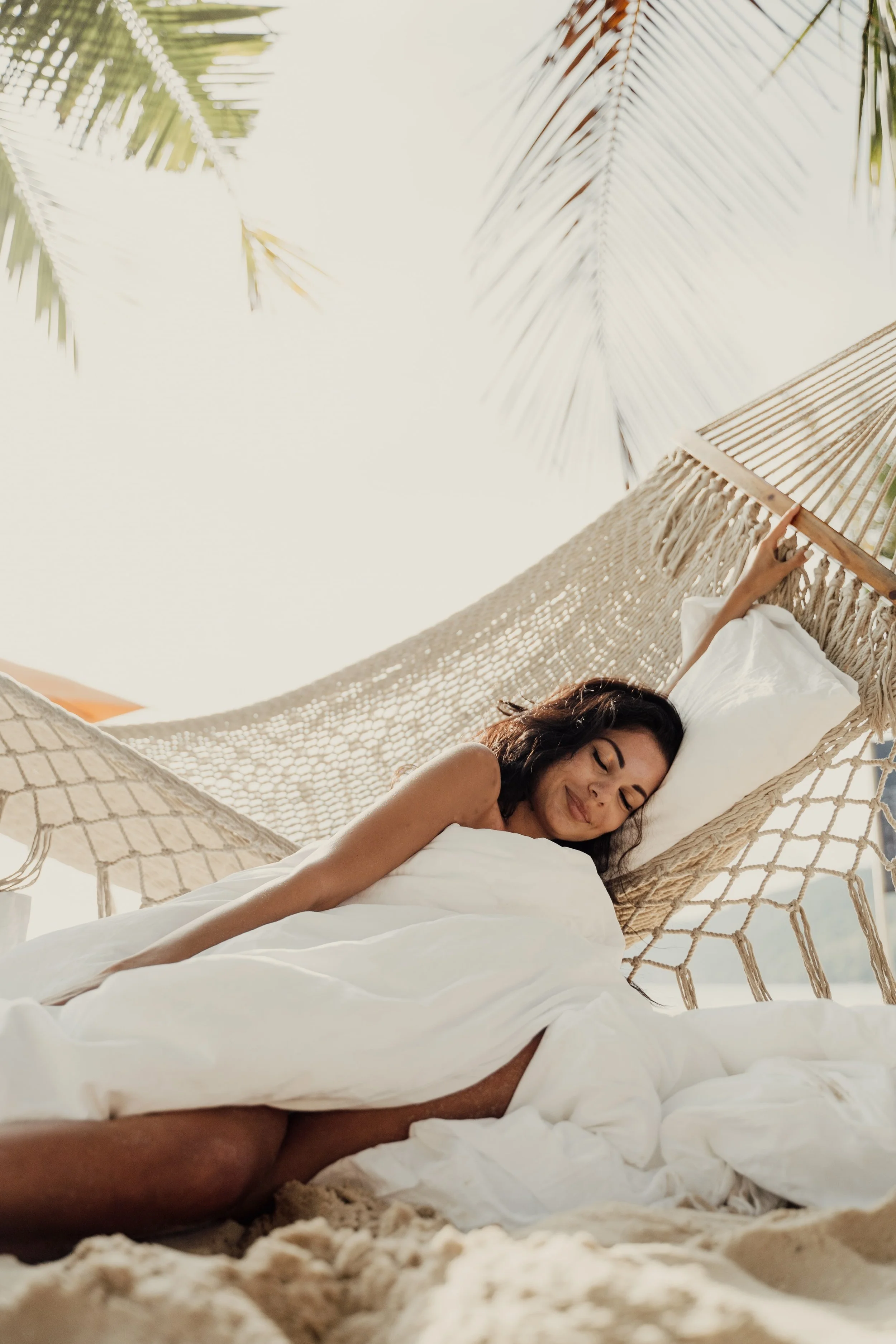 A woman relaxing in a hammock on the beach, smiling with her eyes closed, surrounded by palm trees.