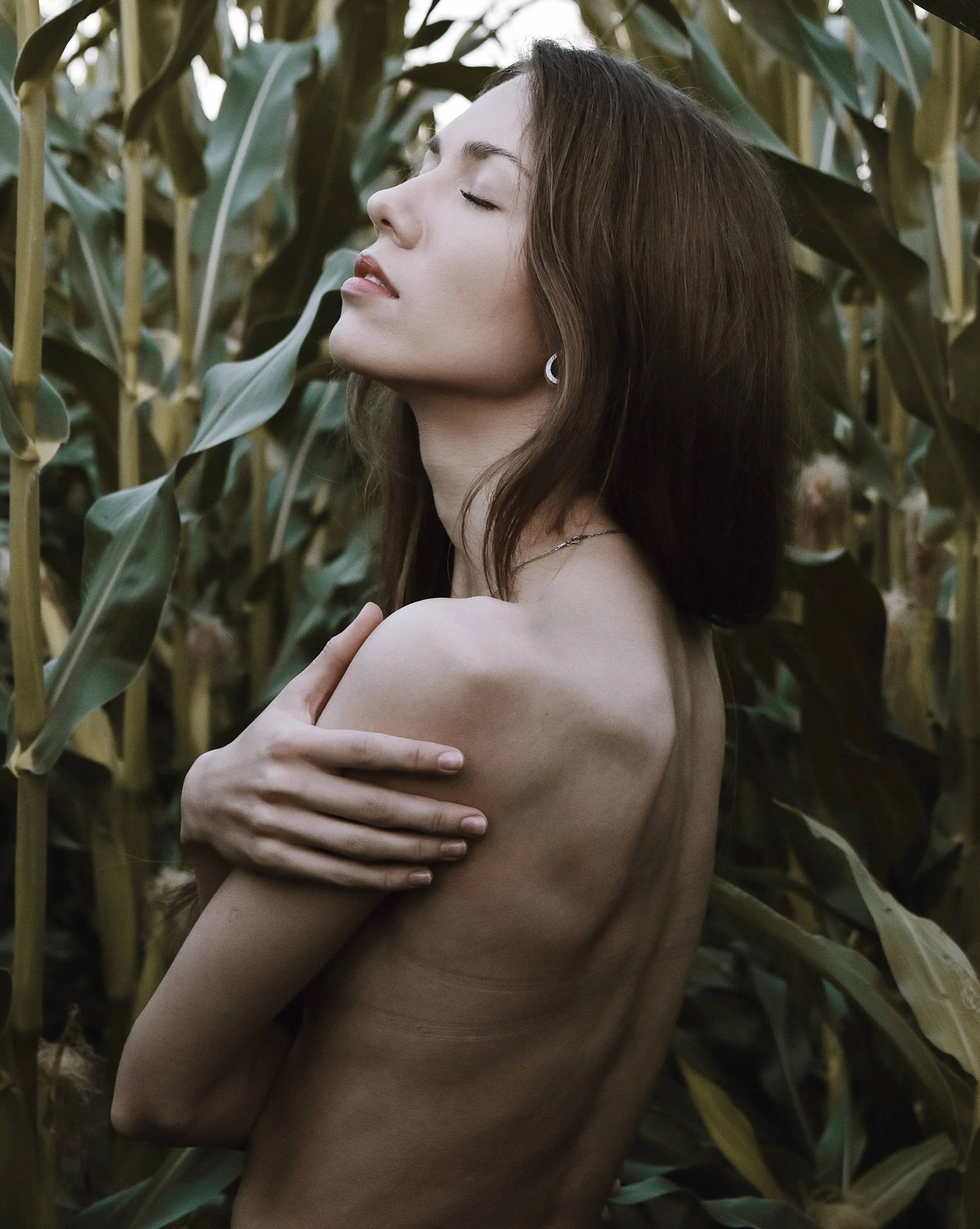 A woman with closed eyes and a serene expression, topless, standing amidst tall corn plants with green leaves. Embracing nature and her skin