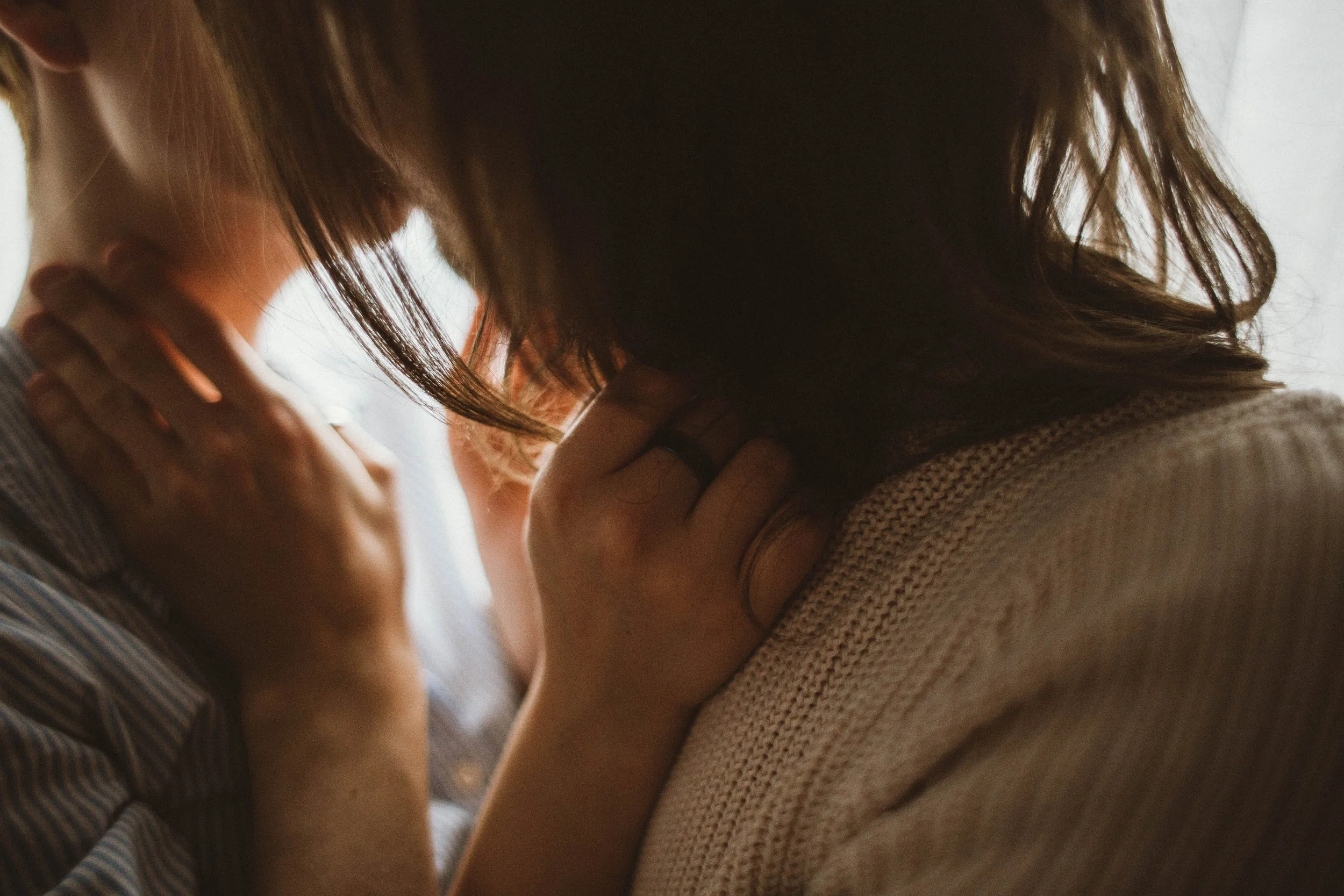 Two women with brown hair gently touching each other's necks while looking down, in an intimate moment indoors with soft natural lighting.