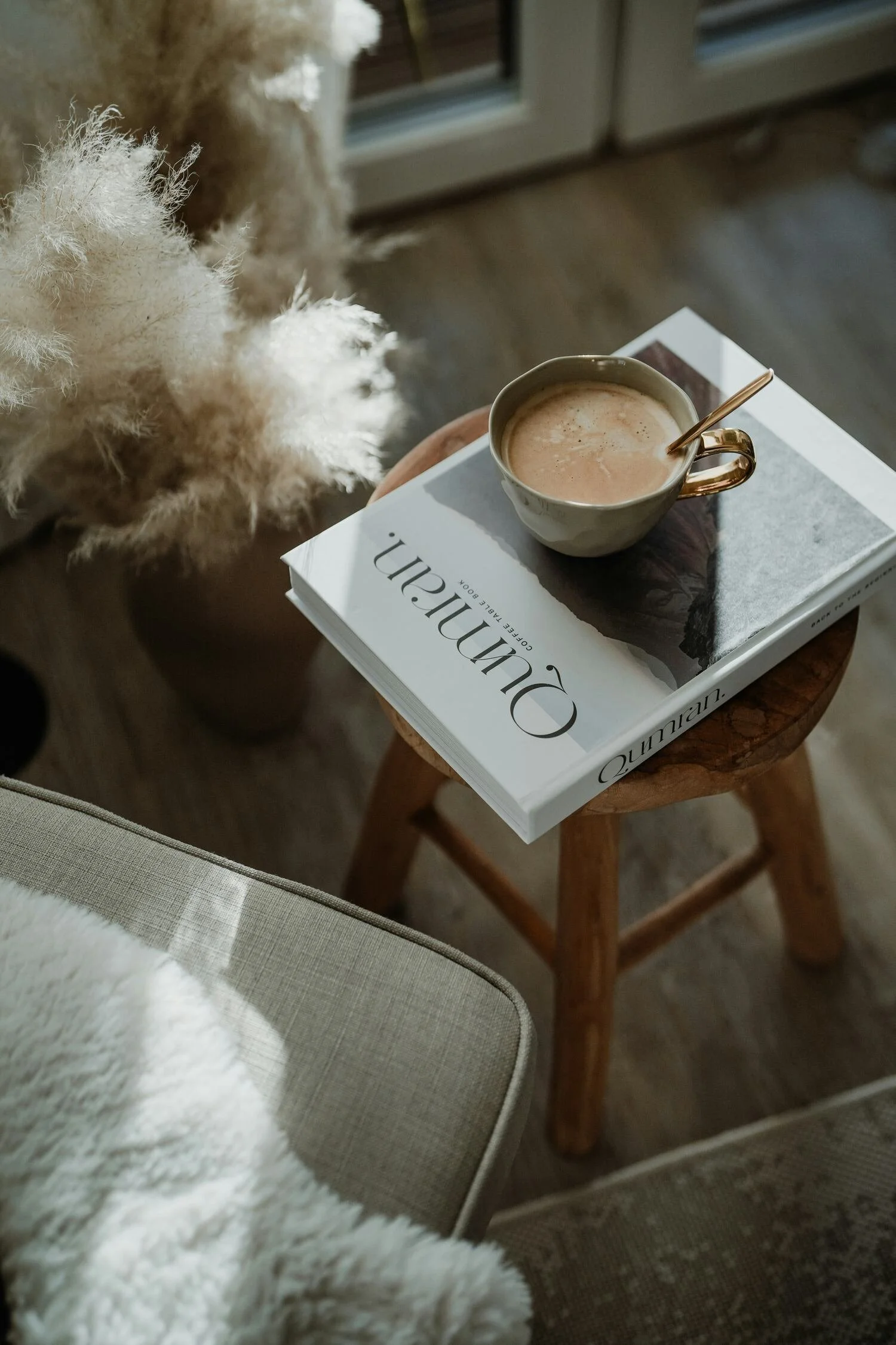 A cup of coffee with a spoon on top, resting on a magazine titled "Quunnian" on a small wooden table near a window. A fluffy white dog is partially visible in the foreground and a potted plant with beige, fluffy foliage is nearby.