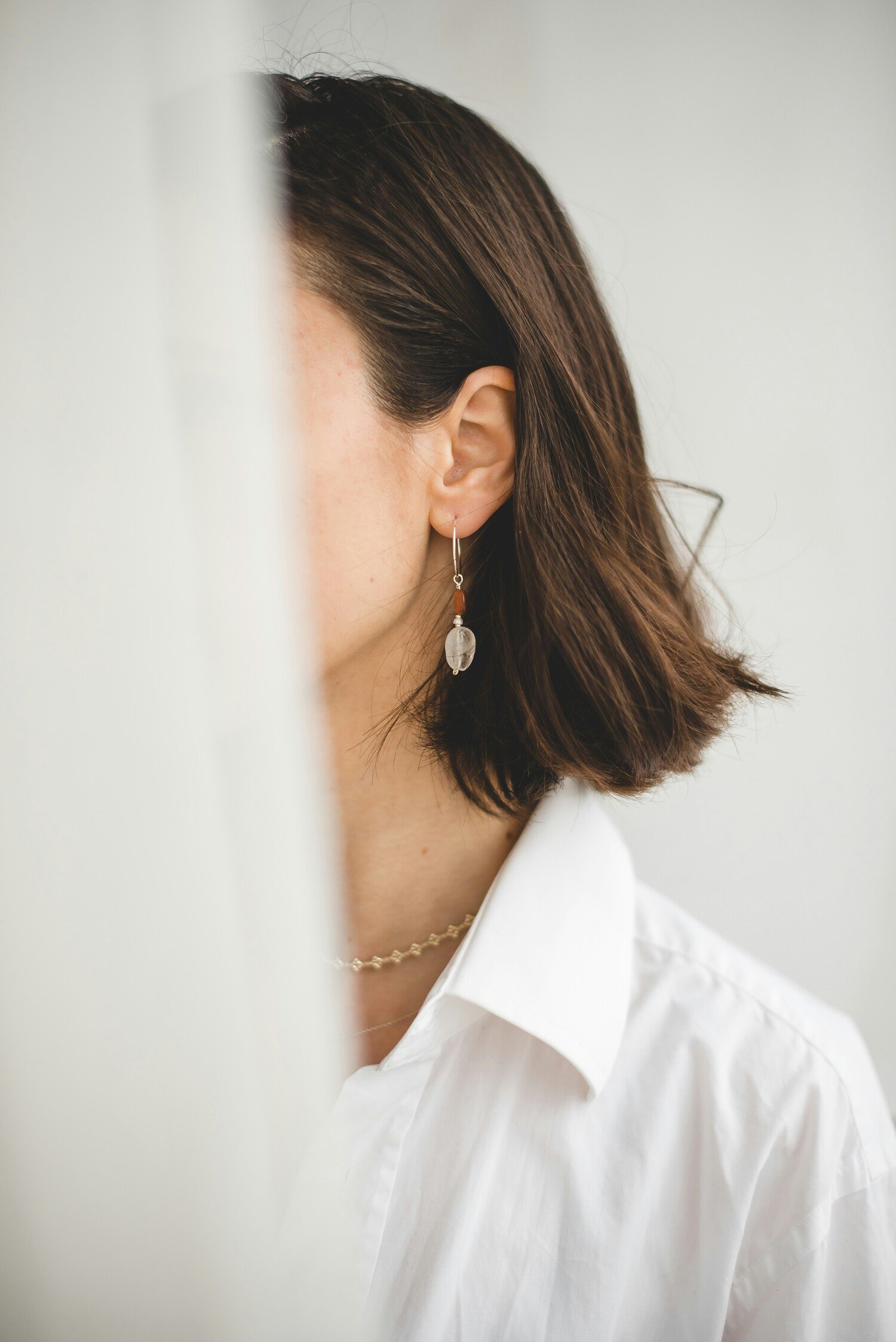 Close-up of a woman with short brown hair wearing earrings and a white shirt, partially hidden behind a white curtain.