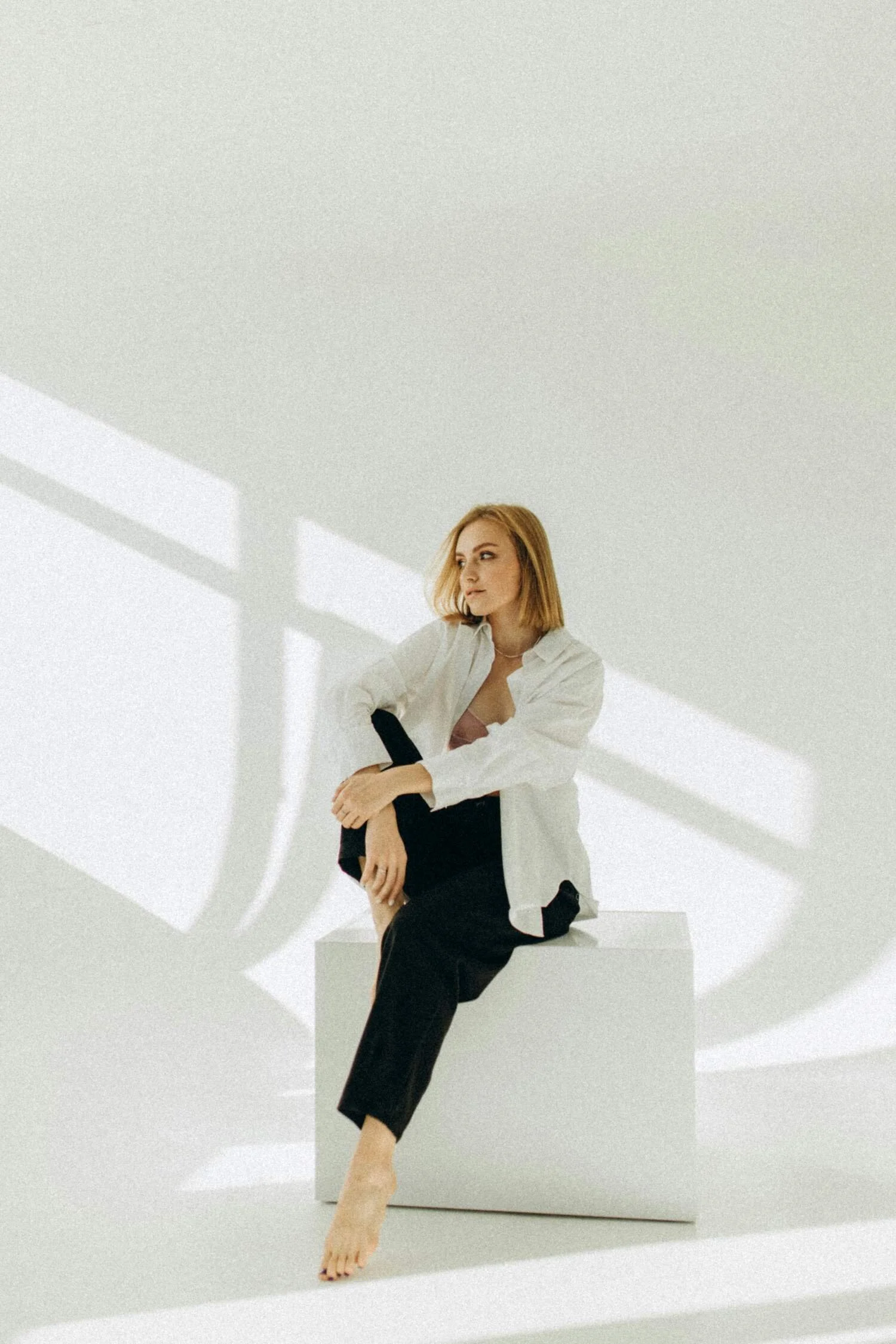 A woman with red hair wearing a white shirt and black pants sitting barefoot on a white box in a minimalist studio with shadows of a bridge or window behind her.