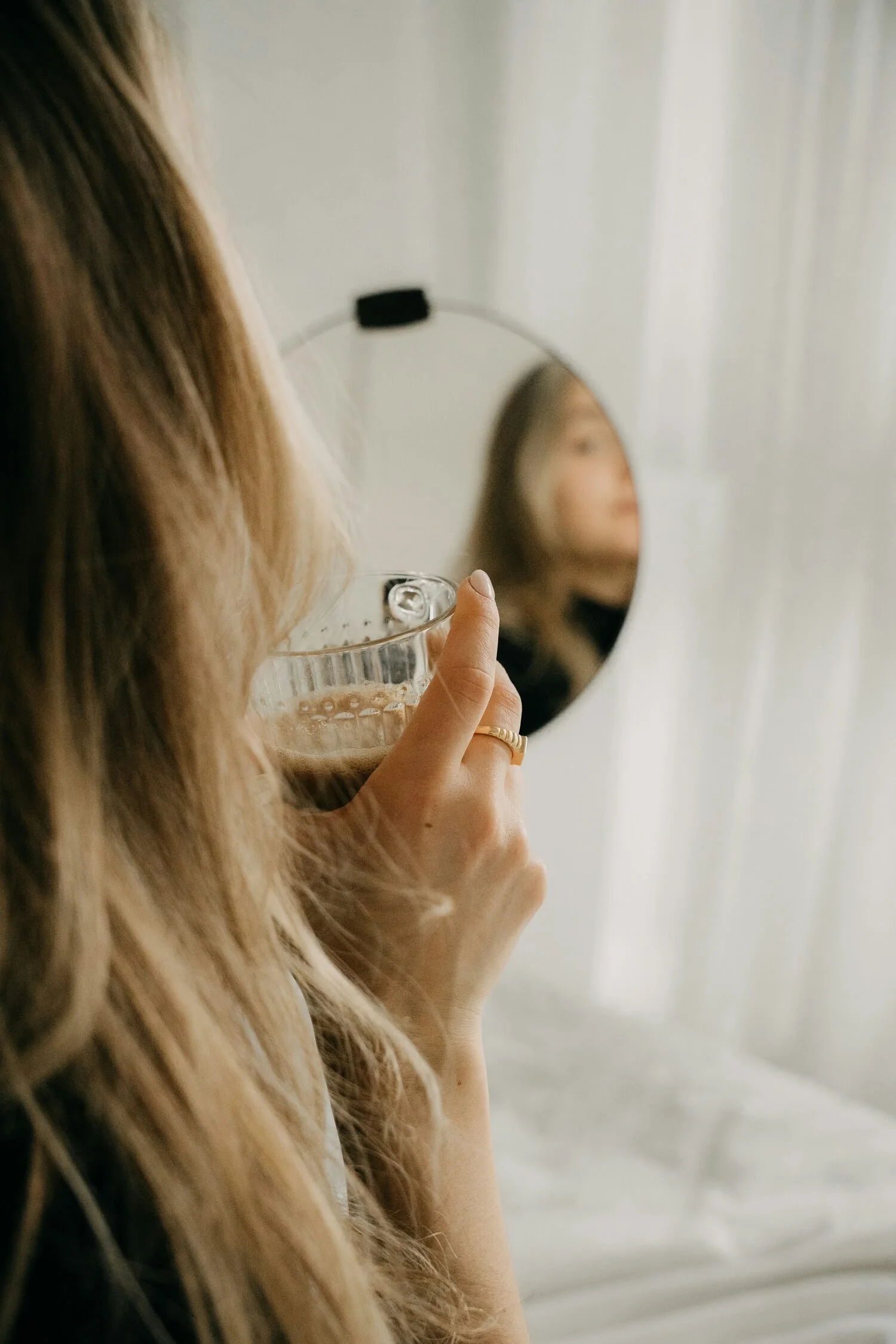 A woman with long blonde hair holding a glass of coffee, looking into a round mirror, with her reflection visible, in a softly lit room with white curtains.
