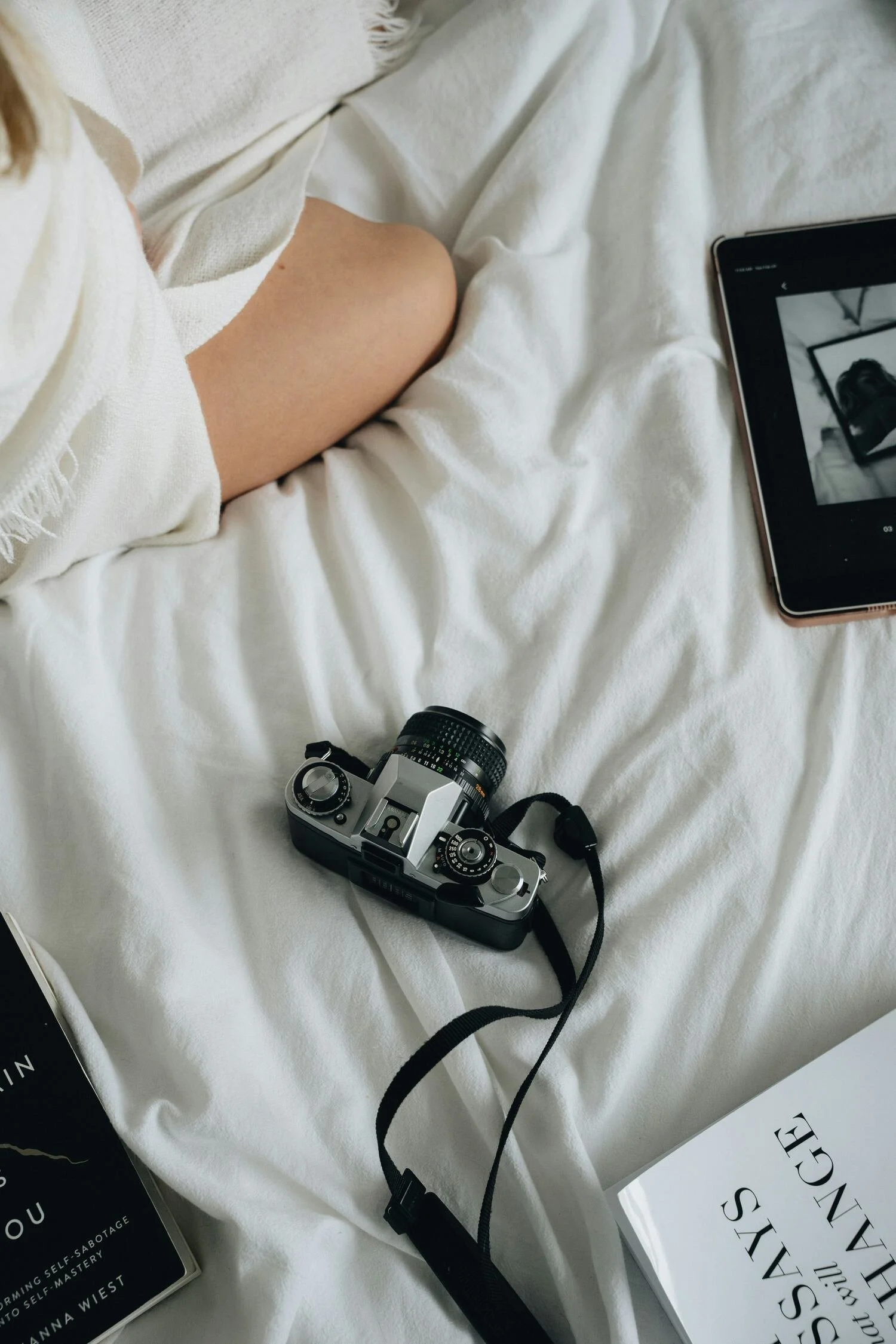 A camera, a tablet showing a black-and-white photo, and a book titled 'The Secrets We Keep' are arranged on a white bed with a person's knee and lap partially visible.