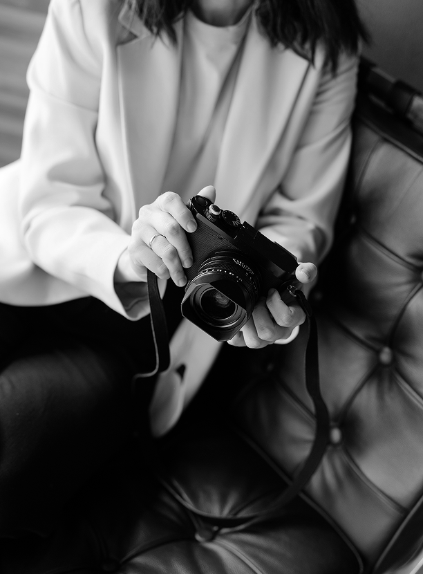 A woman sitting on a leather couch holding a camera in her hands, dressed in a white shirt, with her face partially out of the frame.