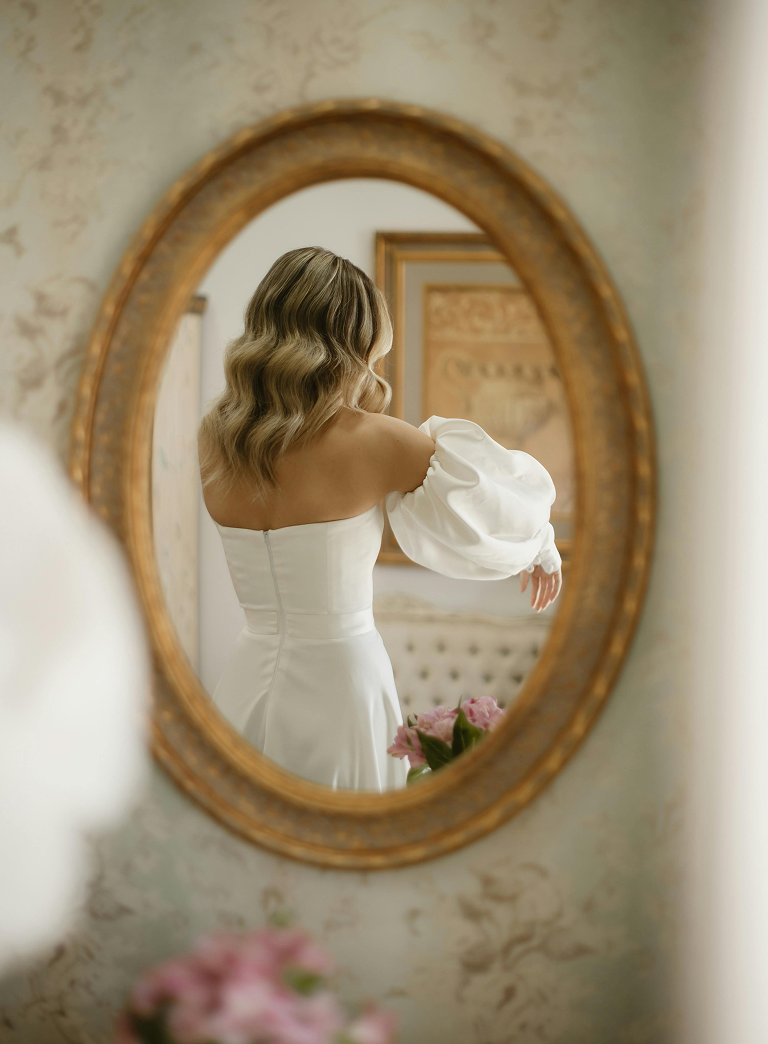 A woman with wavy, blonde hair wearing a white off-the-shoulder dress with puffed sleeves, looking at herself in an oval mirror.