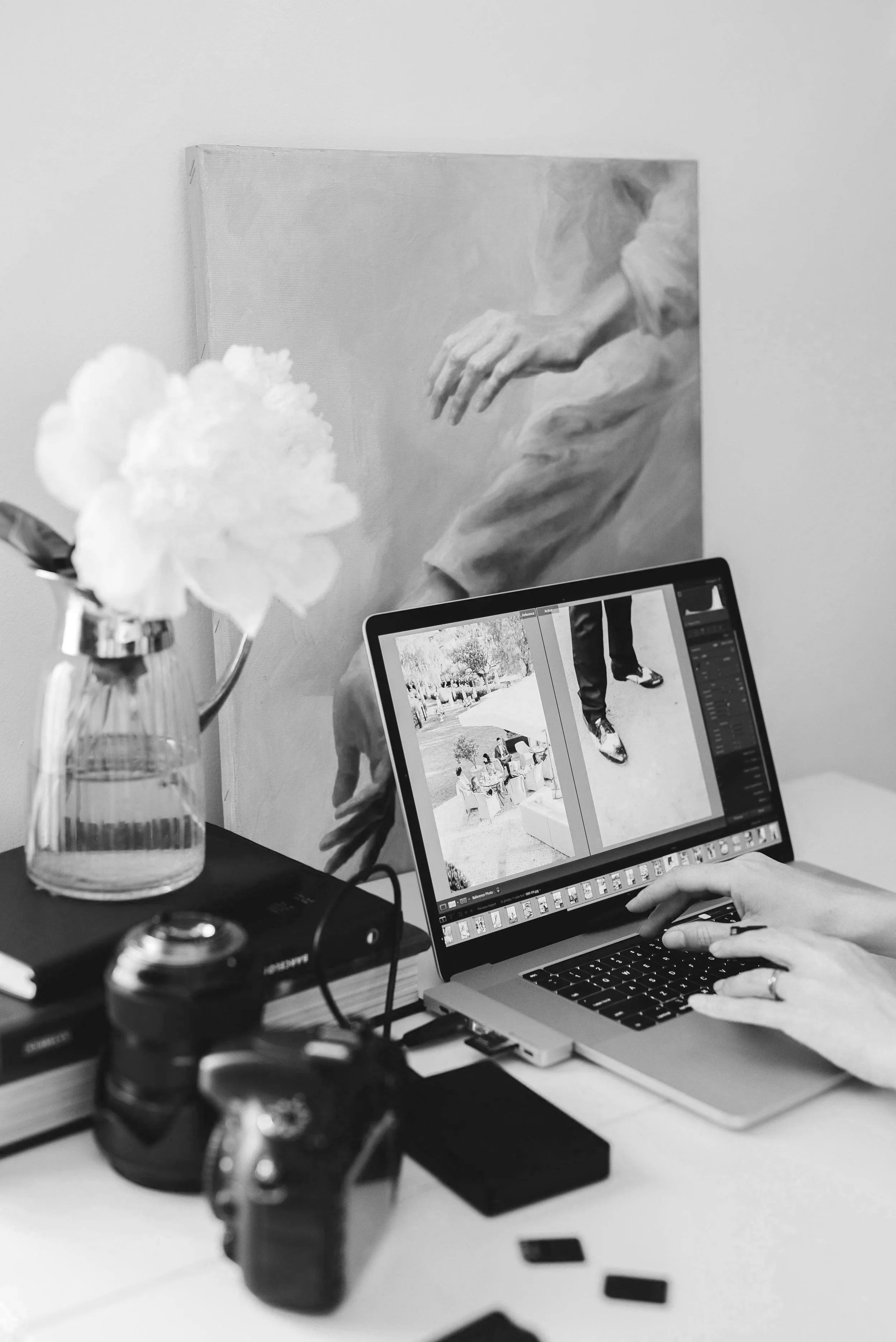 A person editing photographs on a laptop in a workspace with a painting of a hand and arm, a vase with a flower, and camera equipment on the desk.