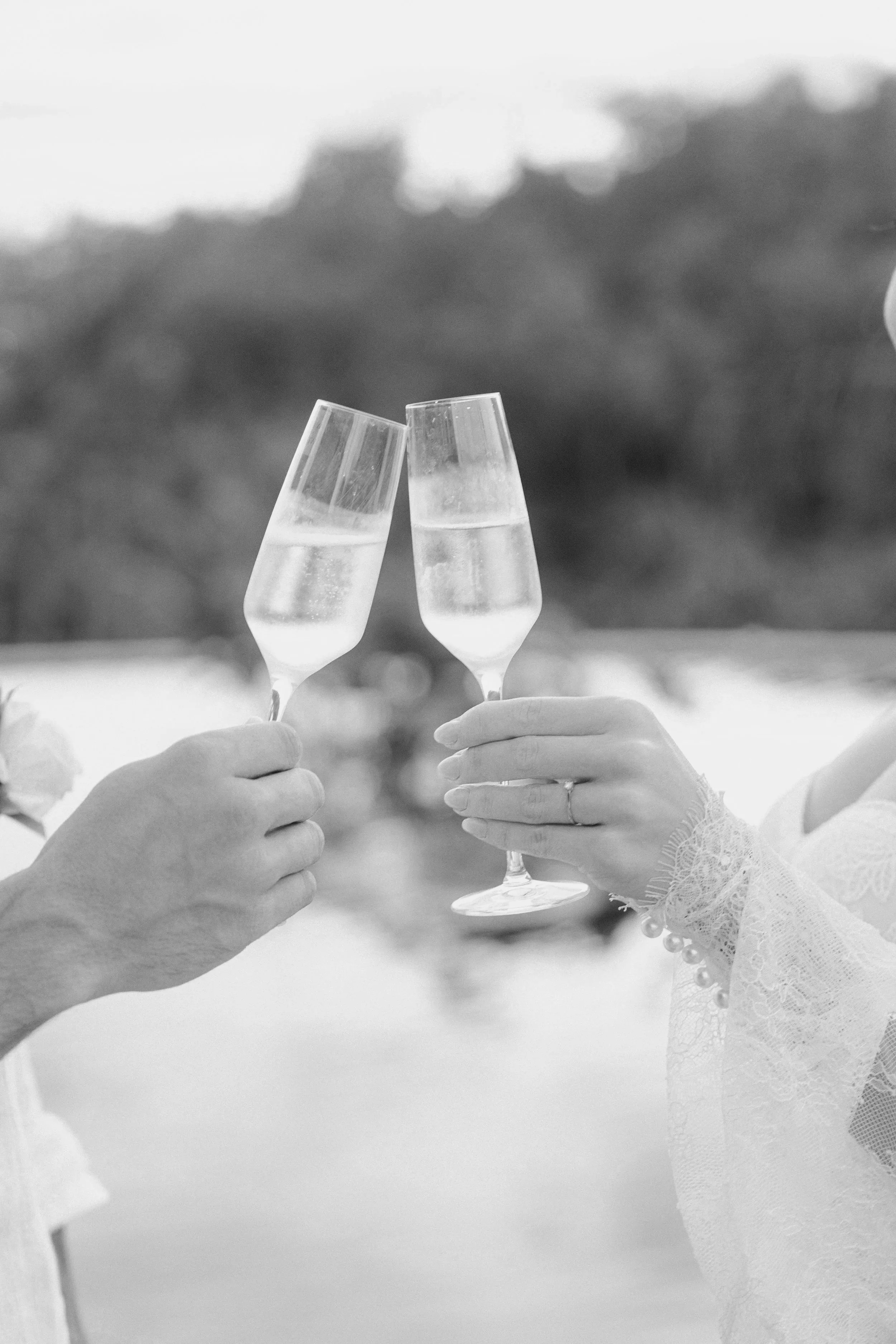 Two people clinking glasses of champagne in a toast, one person's hand with a wedding ring, in black and white.