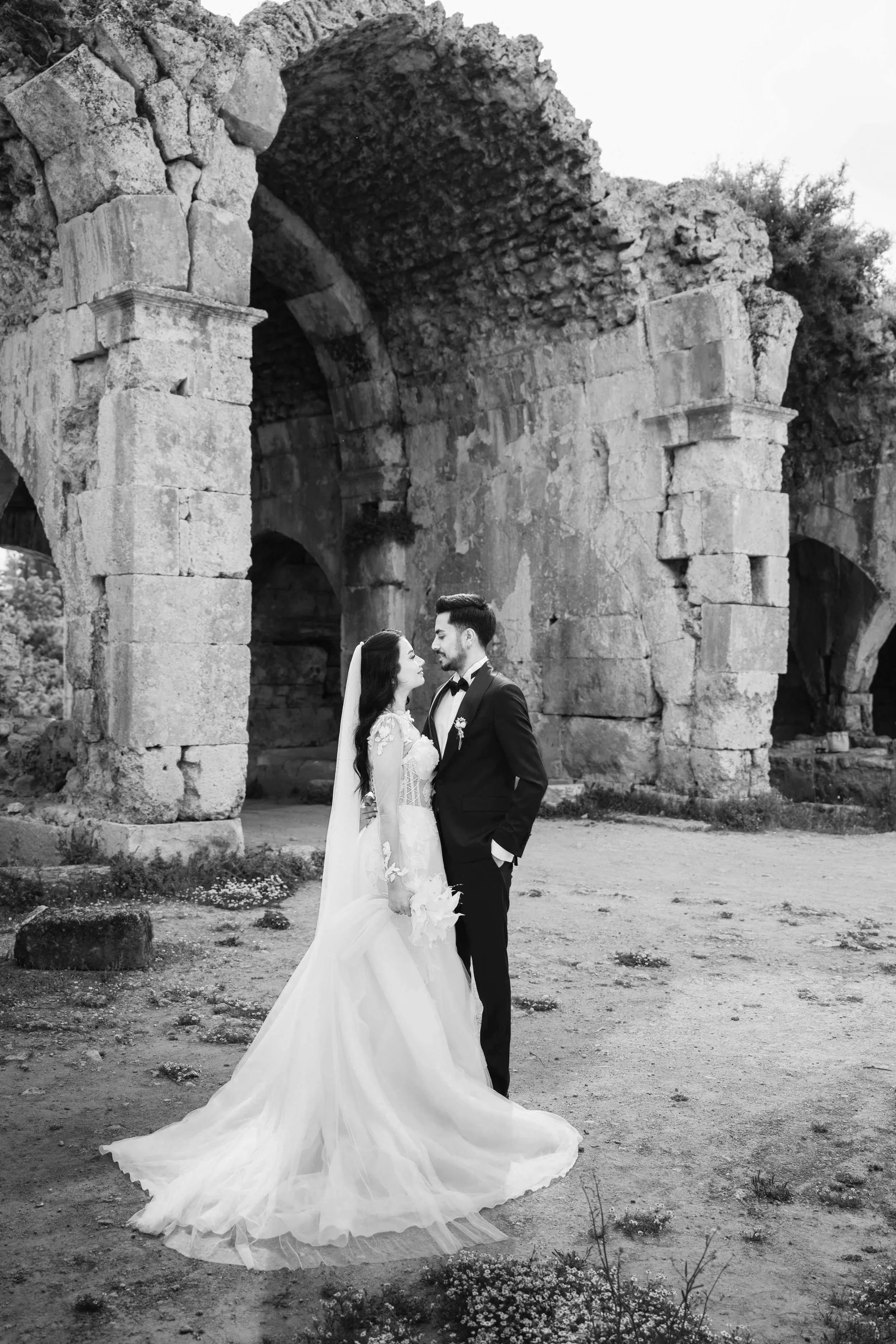 Black and white photo of a bride and groom standing close together in front of ancient stone ruins, looking at each other lovingly.
