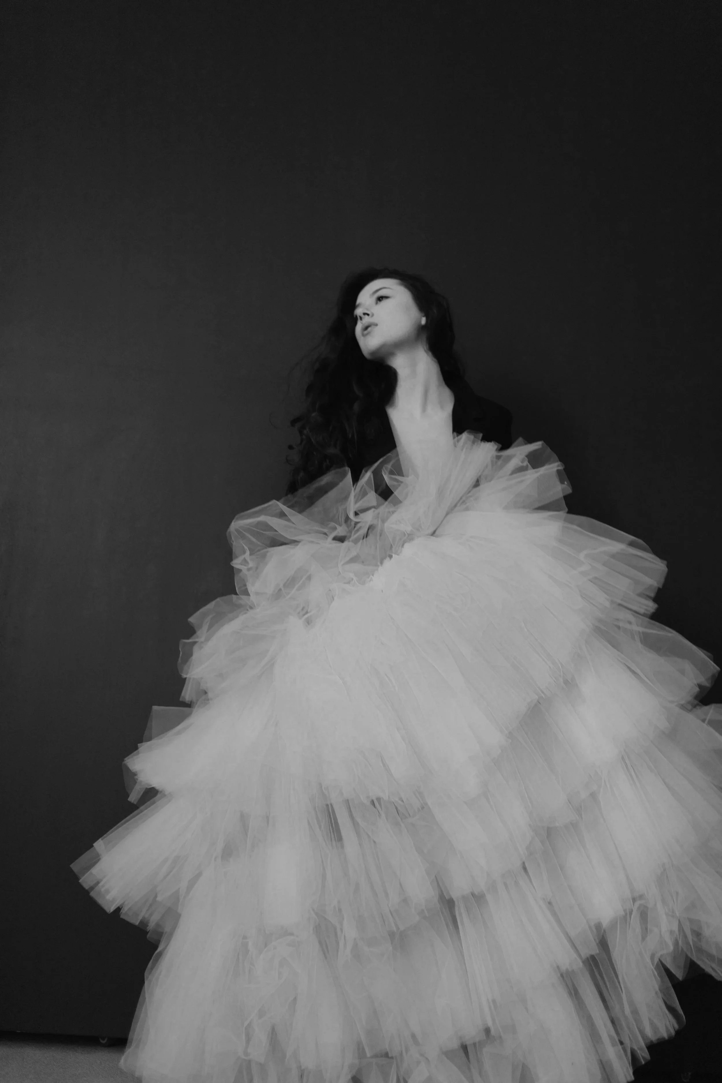 Black and white photo of a woman with long, wavy hair wearing a voluminous, layered tulle dress against a dark background.