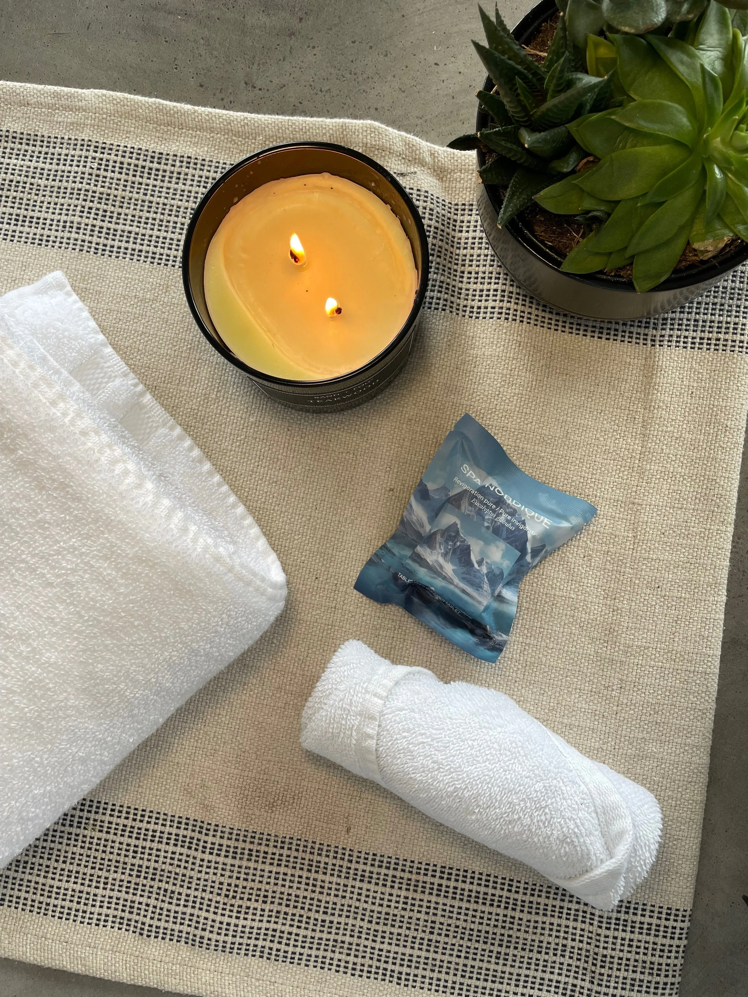 A lit candle in a black container, a small potted succulent plant, a folded white towel, a rolled white towel, a small box of SeaNouDUE bath salts, and a white linen mat on a beige fabric surface.